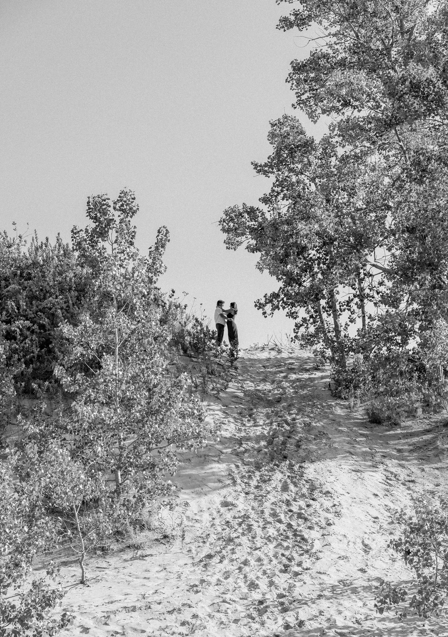 Black and white photo of two people standing on sandy dunes surrounded by lush trees under a clear sky.
