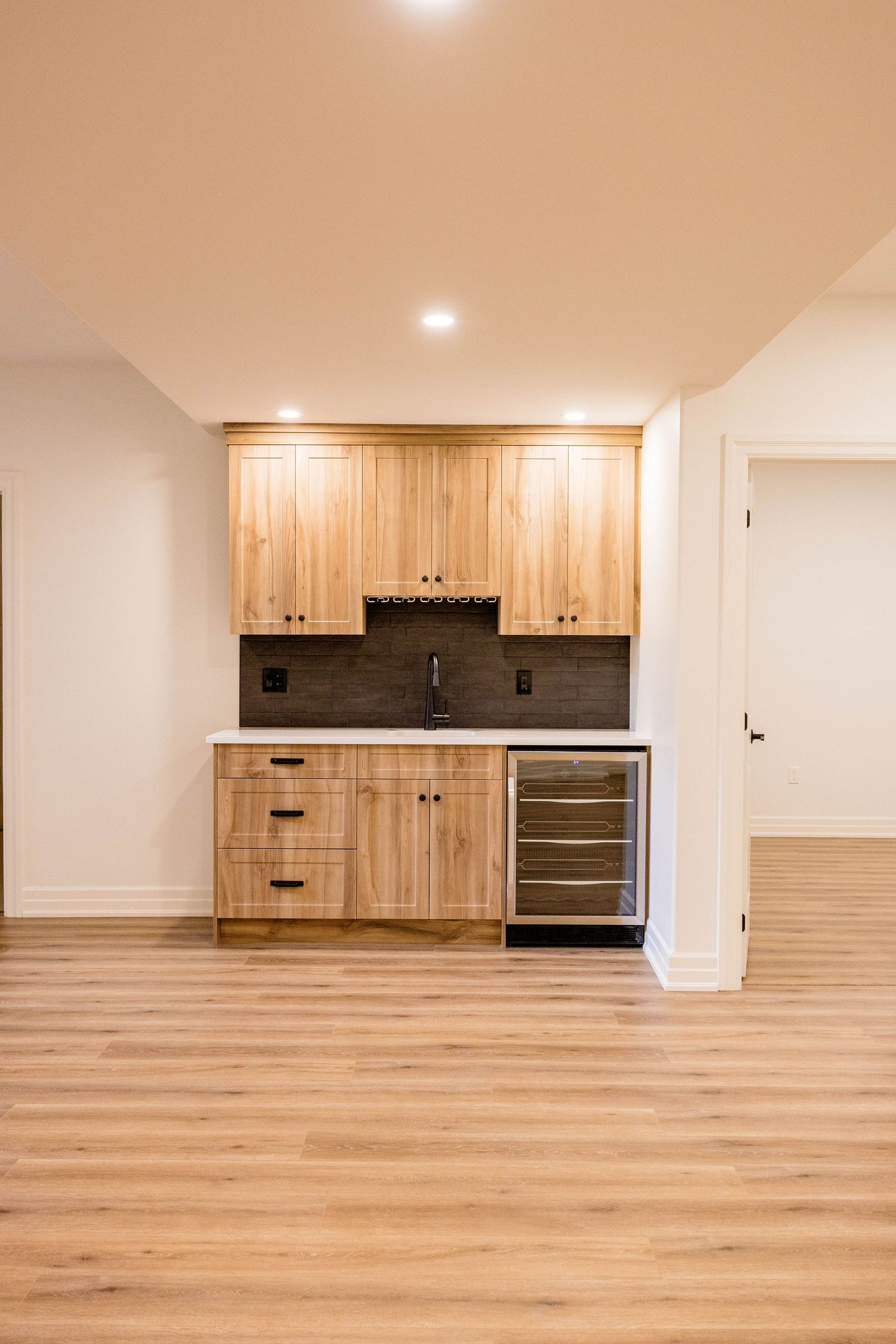 Modern wet bar with light wood cabinets, dark tile backsplash, wine fridge, and hardwood floors in a finished basement.