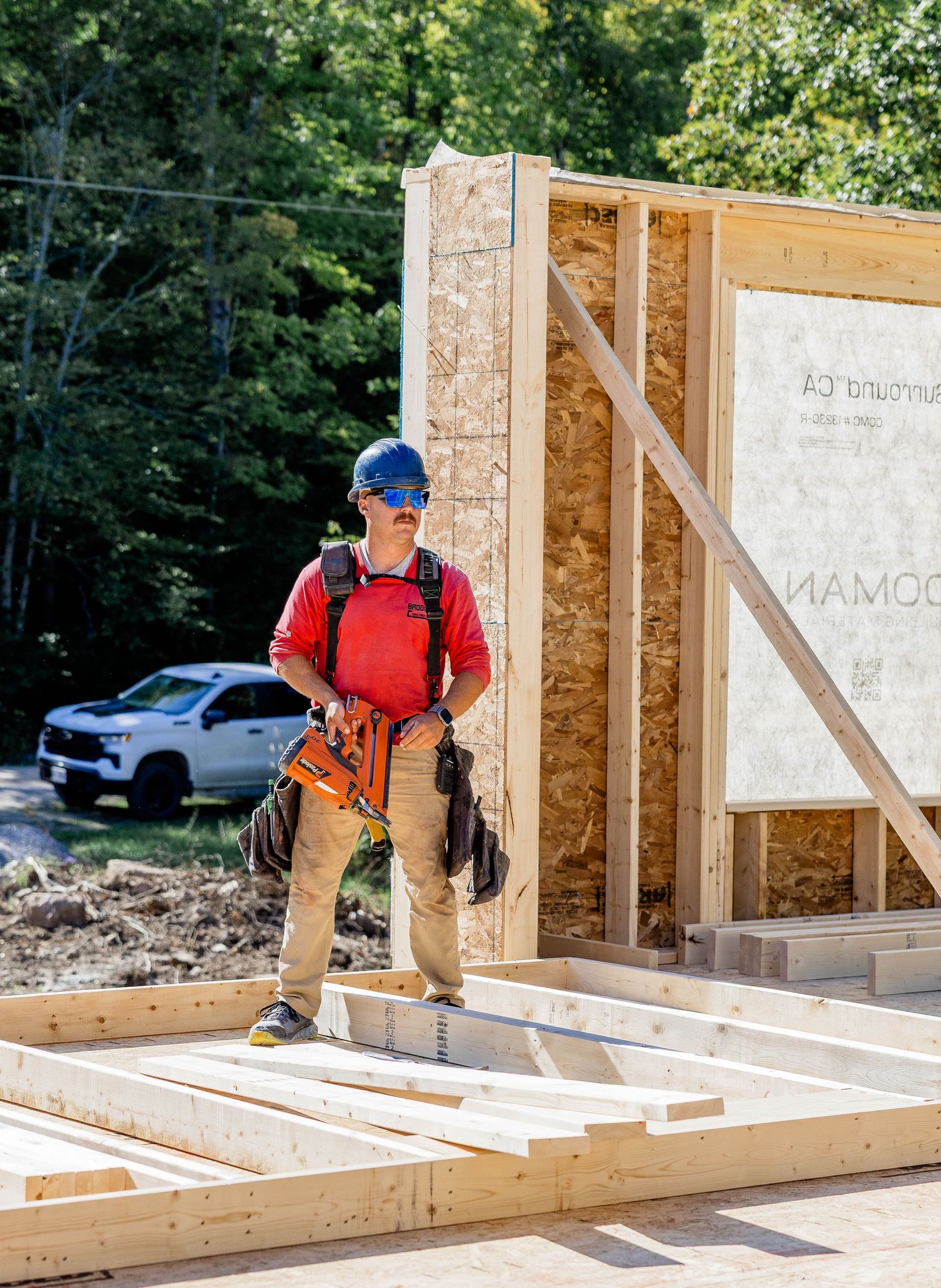 Construction worker in red shirt and blue hard hat holds nail gun on a new home framing site with wood panels.