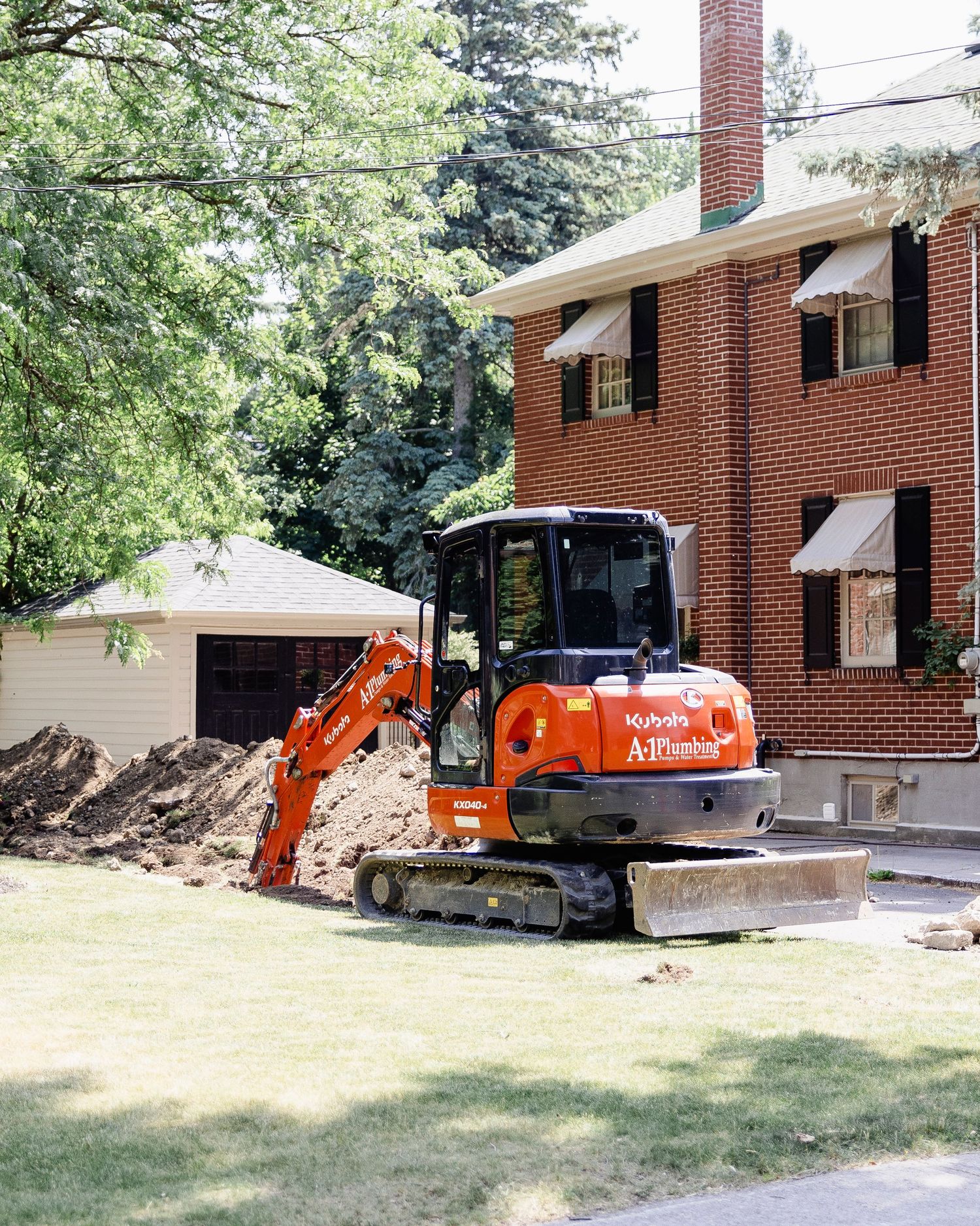 Orange Kubota mini excavator digging near brick residential home with pile of dirt on lawn.