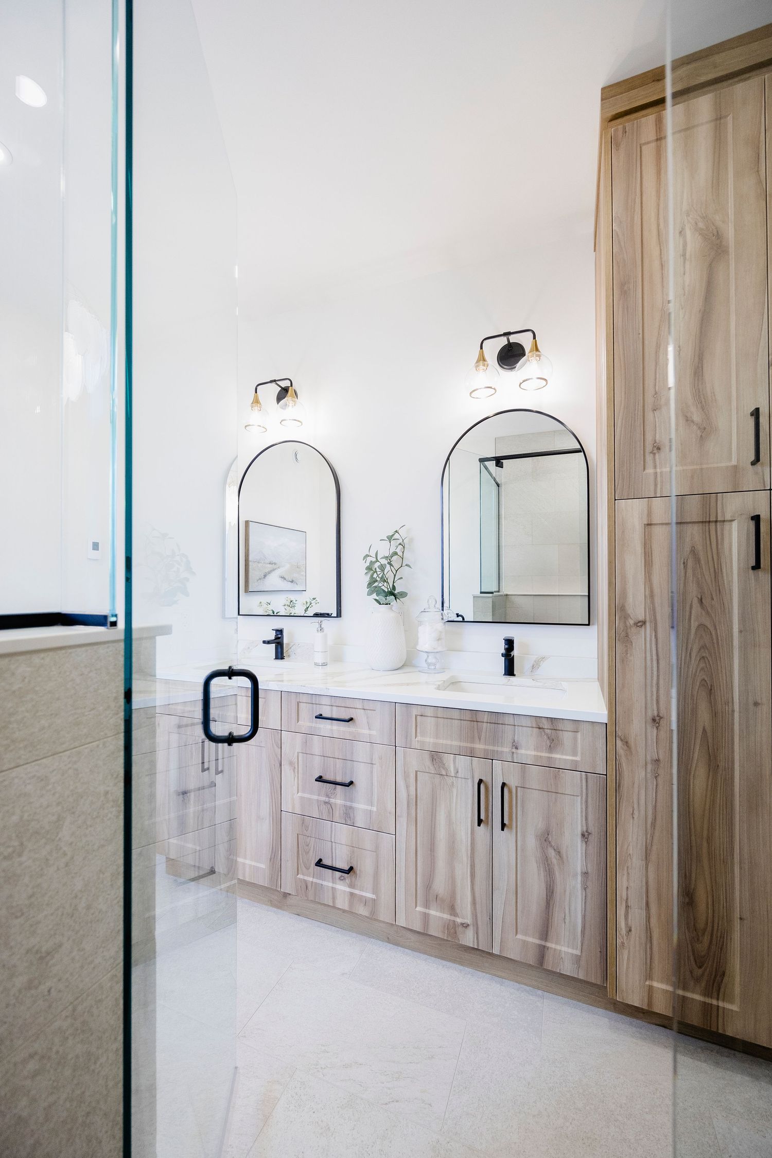 Modern double vanity bathroom with light wood cabinets, arched mirrors, black fixtures, and white countertops.