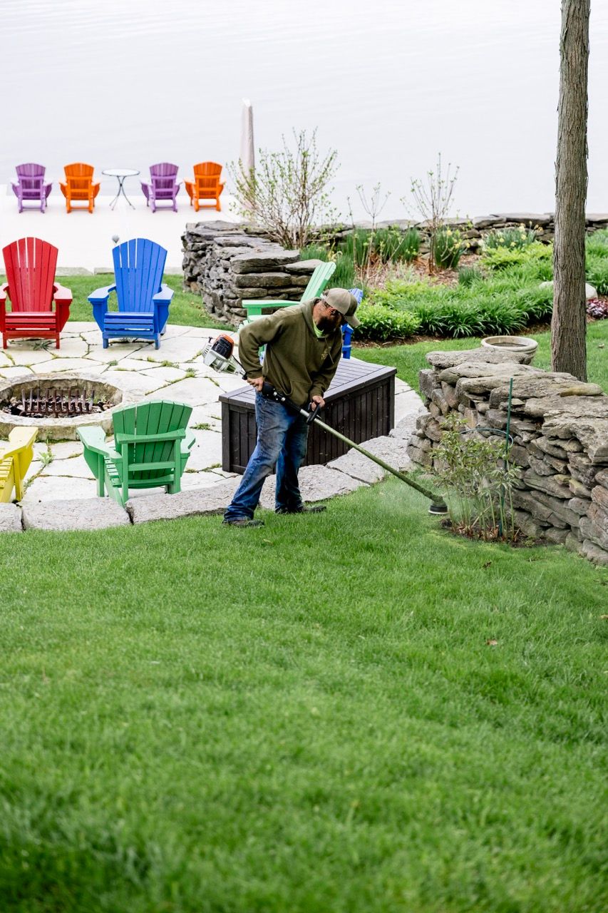 Man edging lush green lawn near colorful Adirondack chairs and stone fire pit patio area.