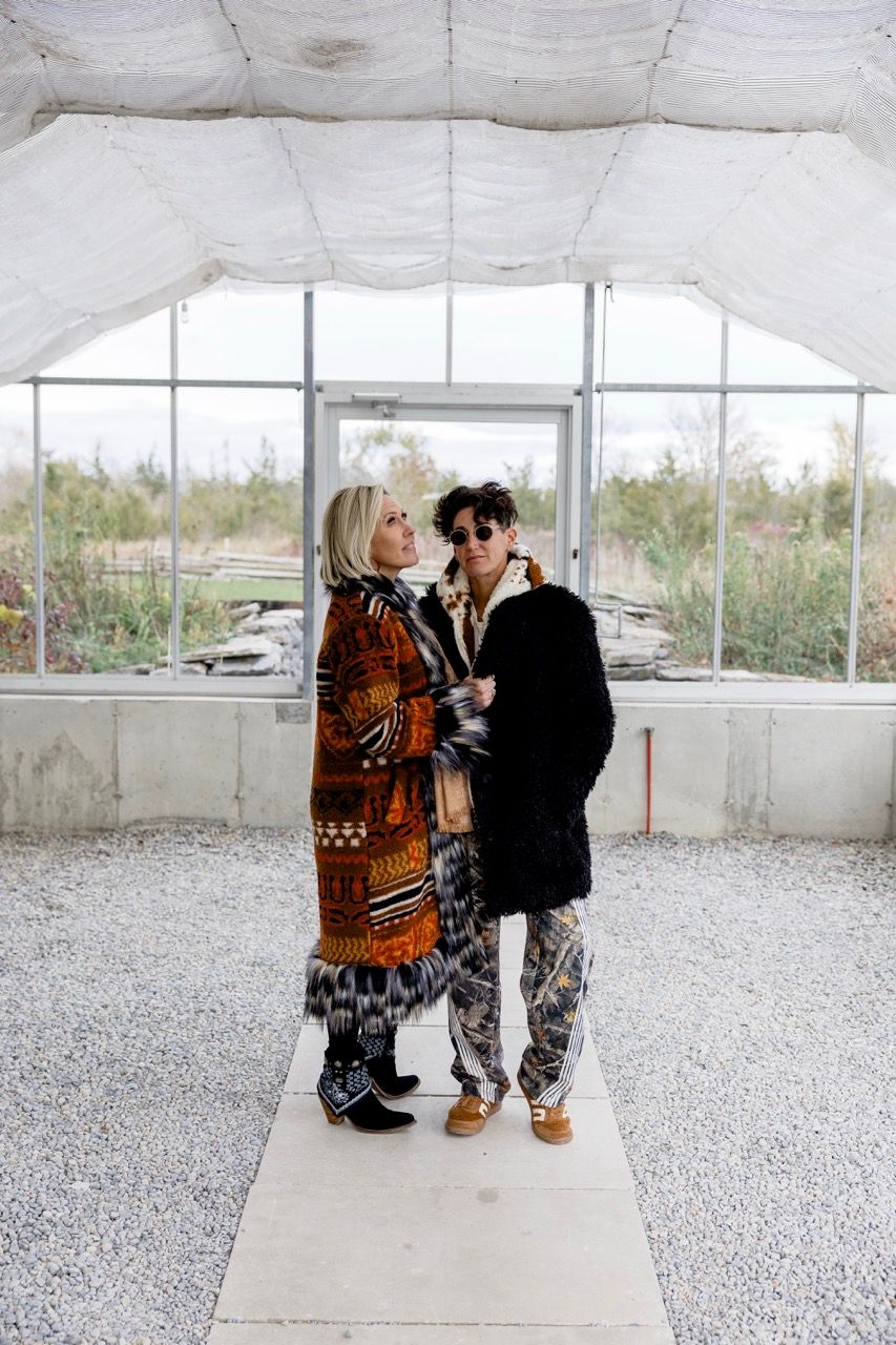 Two people in eclectic outfits stand together inside an unfinished greenhouse with gravel flooring.