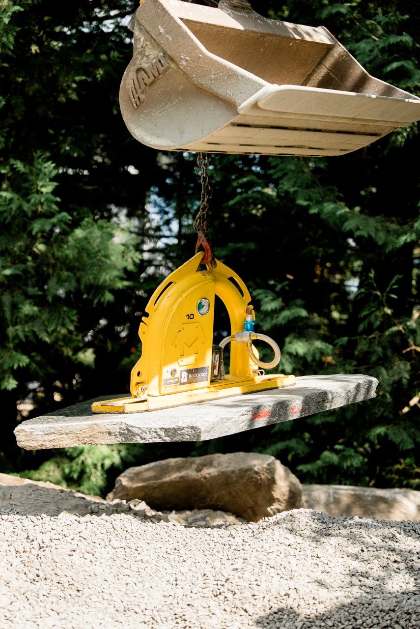 Yellow vacuum stone lifter attachment on excavator bucket lifting flat granite slab outdoors.