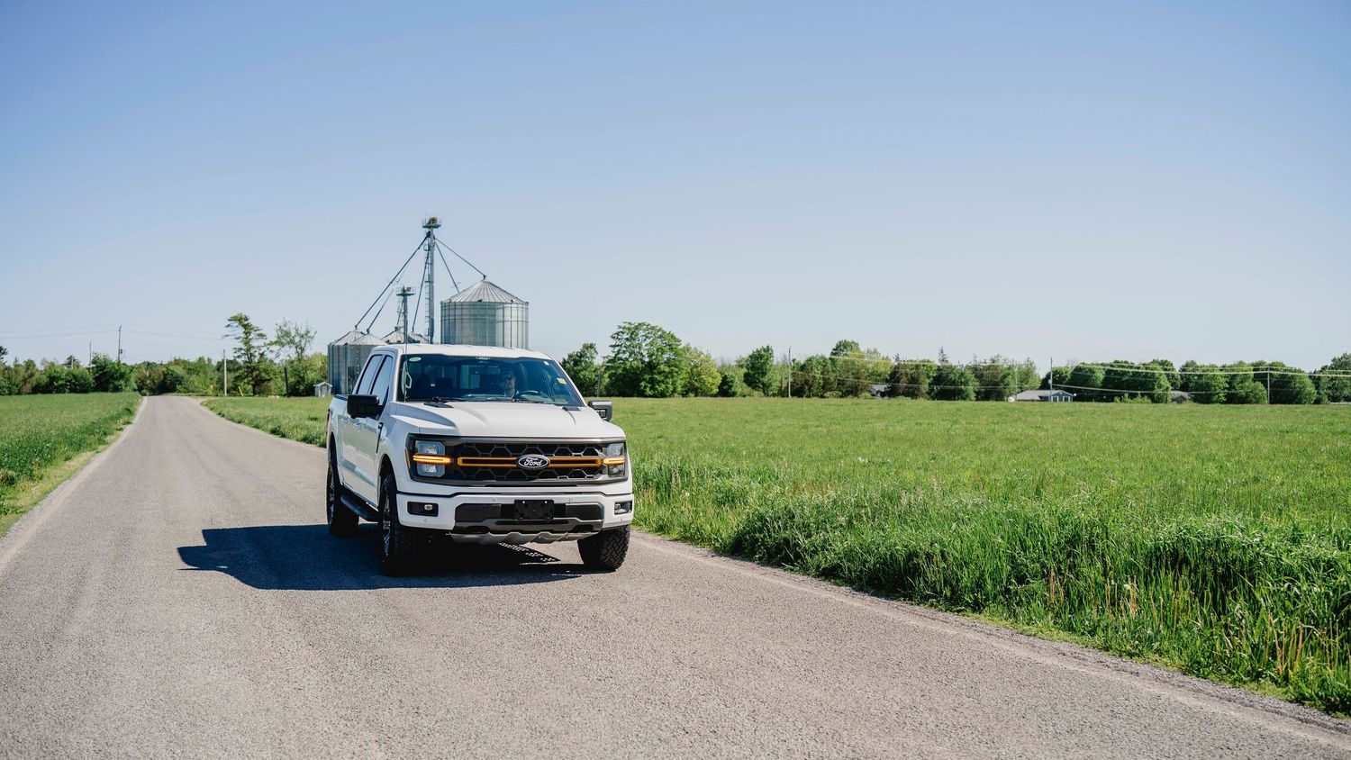 White Chevrolet truck on rural road flanked by green fields and grain storage silos.