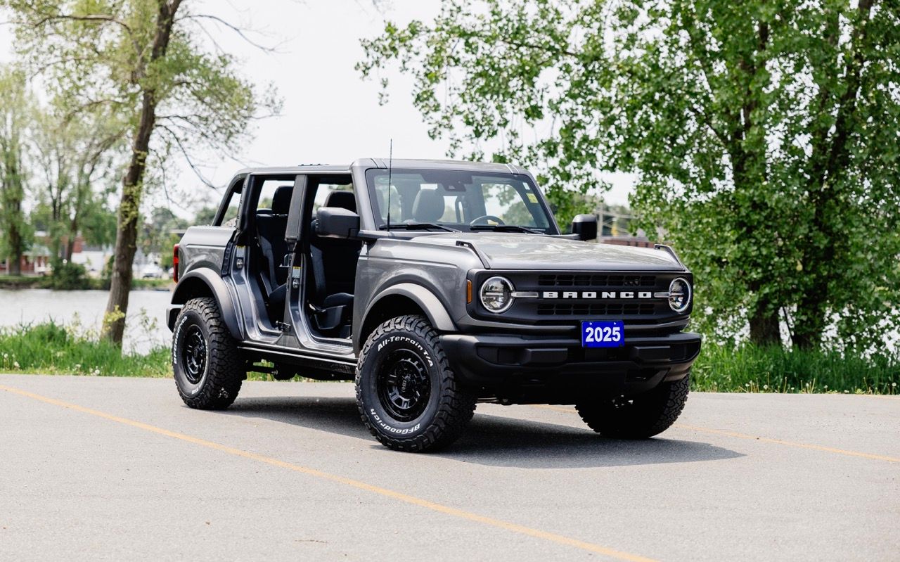 Black 2025 Ford Bronco four-door SUV with no doors parked on a paved lot near green trees.
