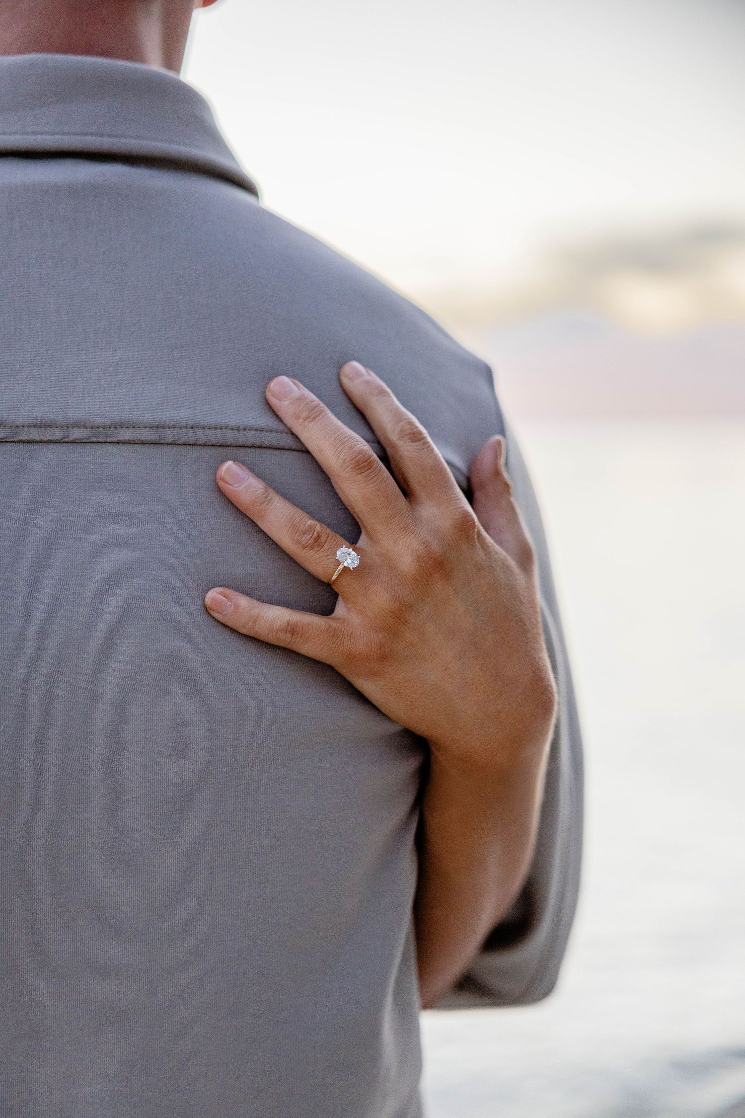 A close-up of a woman's hand with a diamond engagement ring resting on a man's gray shirt shoulder at sunset.
