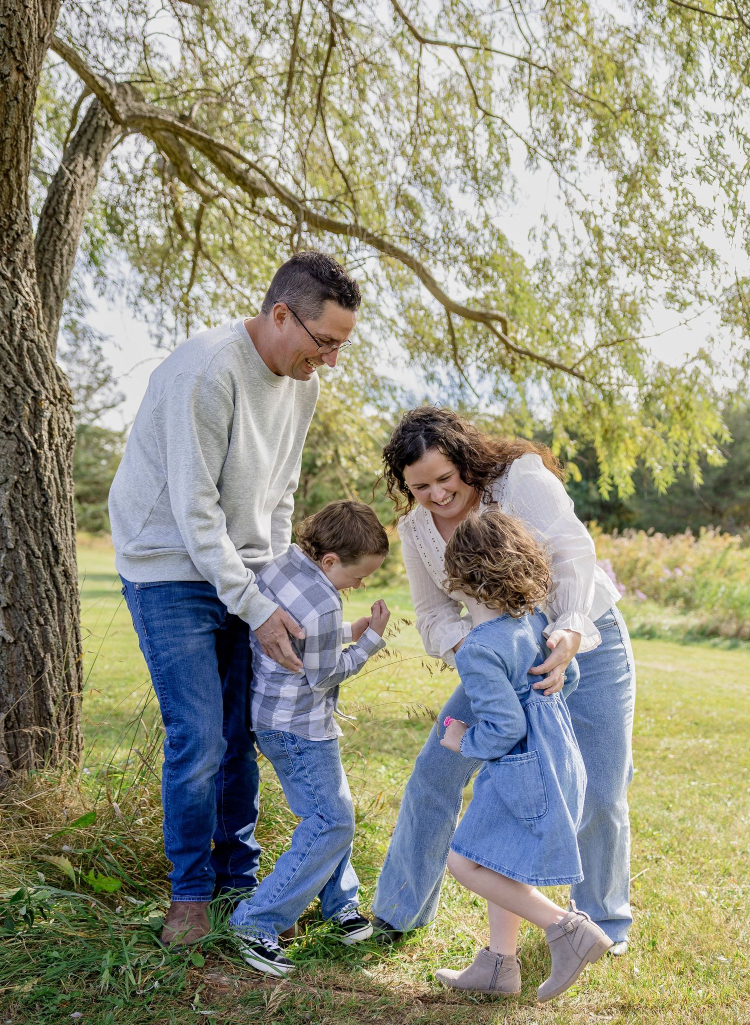 Family of four laughing and playing together under a willow tree in a green park on a sunny day.