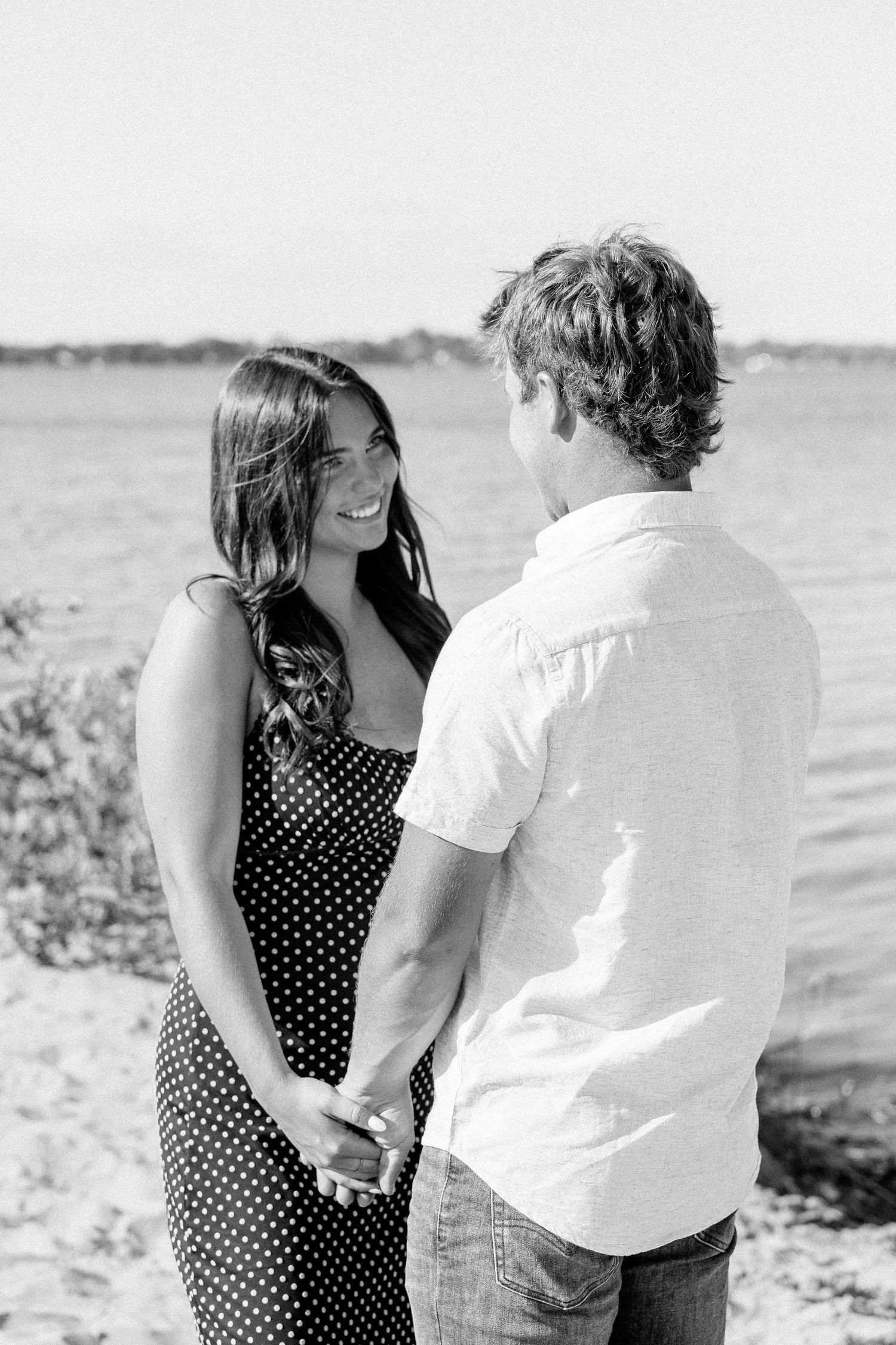 Black and white photo of a couple holding hands and smiling at each other on a sunny beach.