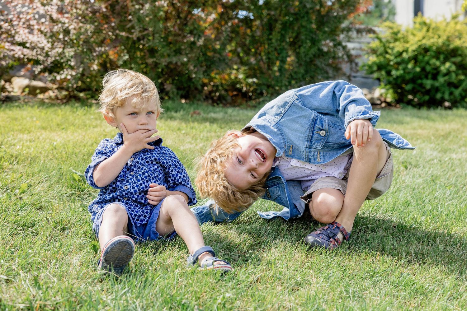 Two young boys playing on green grass outdoors, one sitting and one leaning over smiling, on a sunny day.
