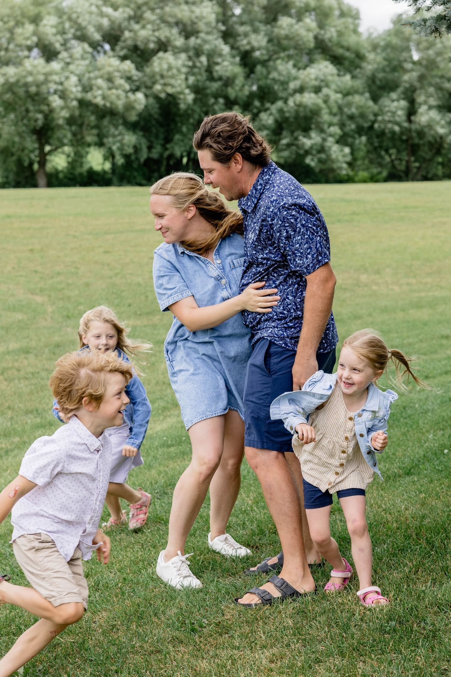 Family with four young children playing and hugging outdoors on a green lawn with trees in the background.