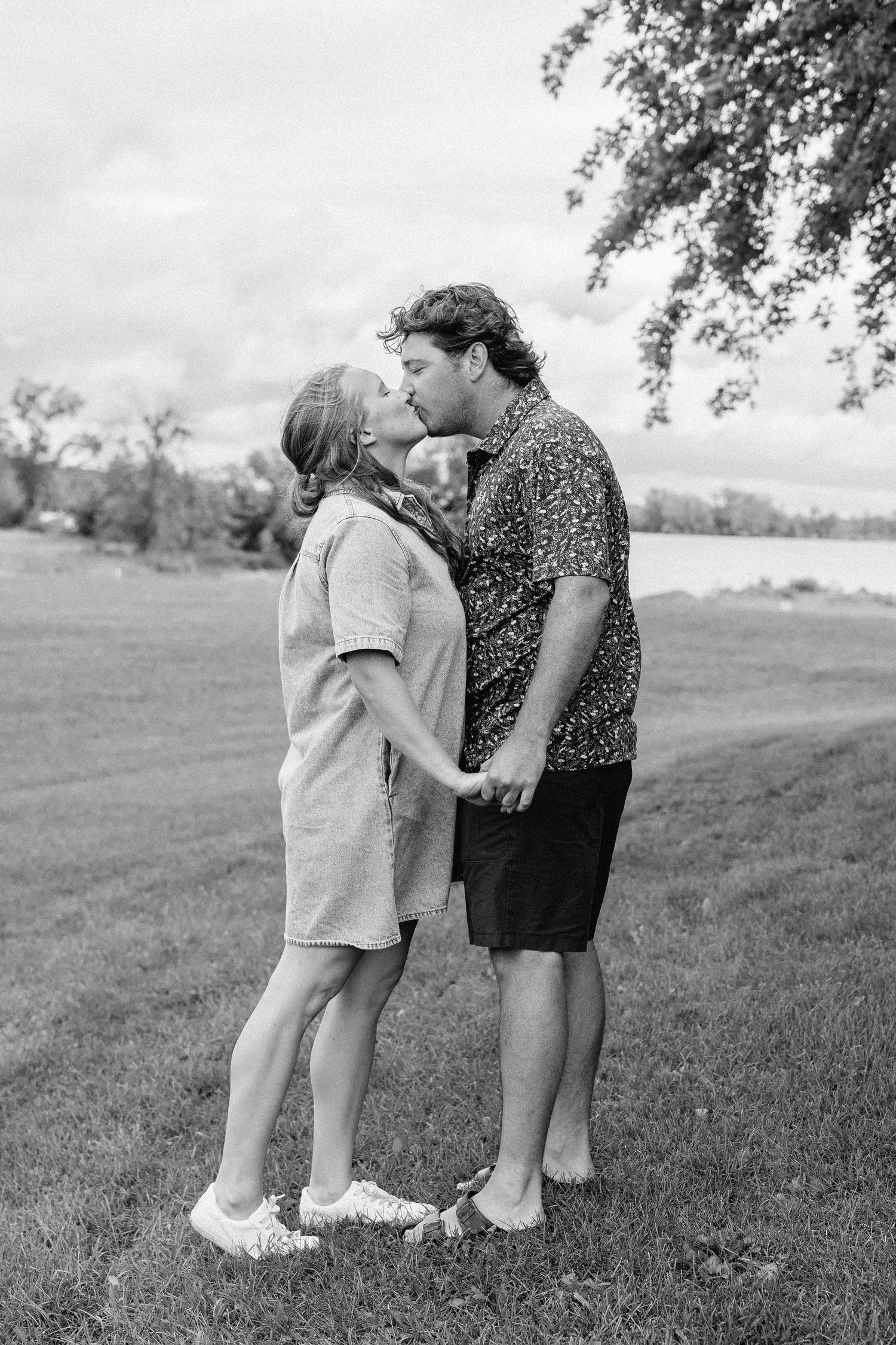 A couple shares a romantic kiss while holding hands in a grassy park, captured in black and white photography.