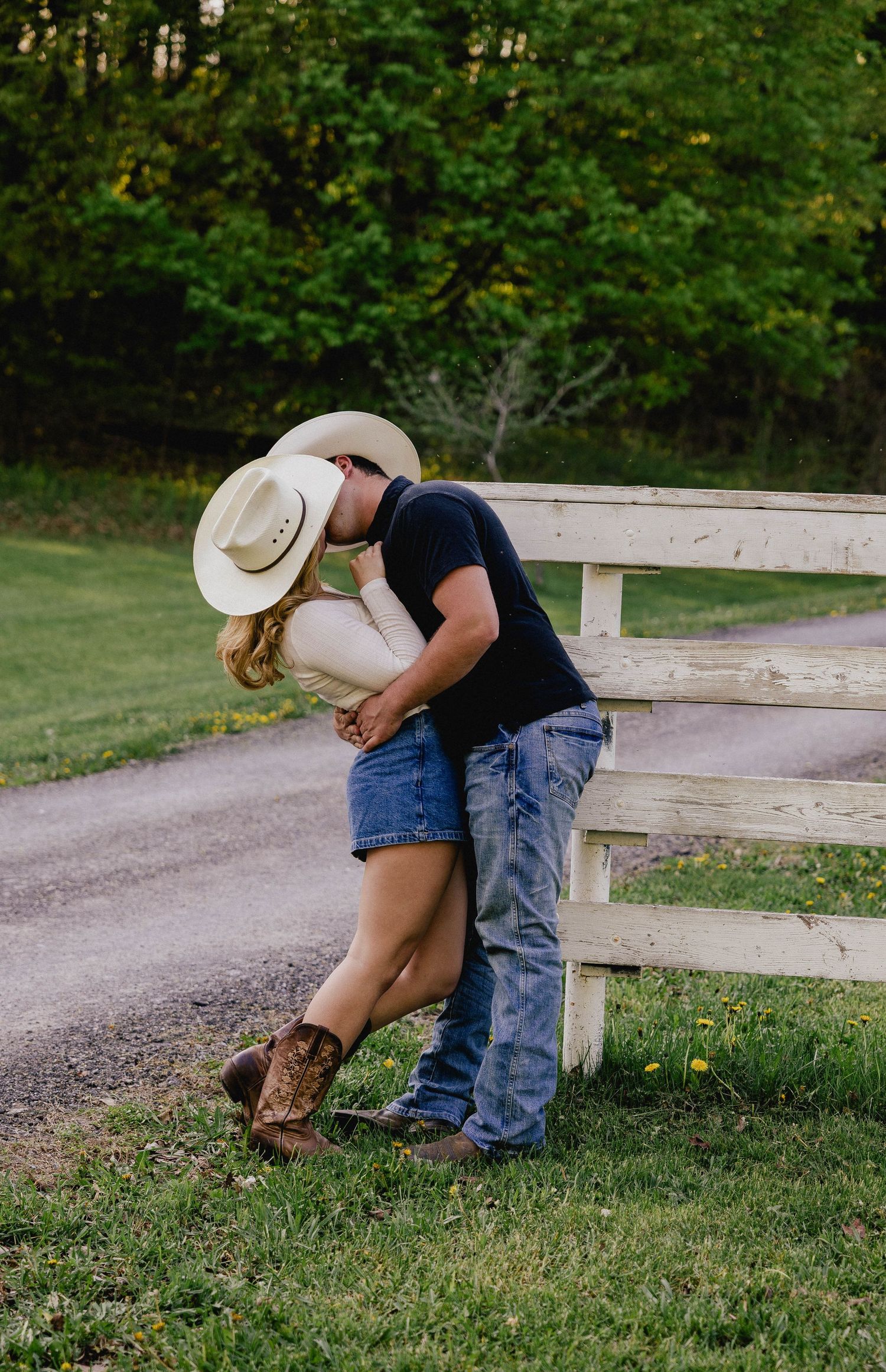 Romantic couple embracing by rustic white fence, woman in cowboy hat and denim skirt with cowboy boots.