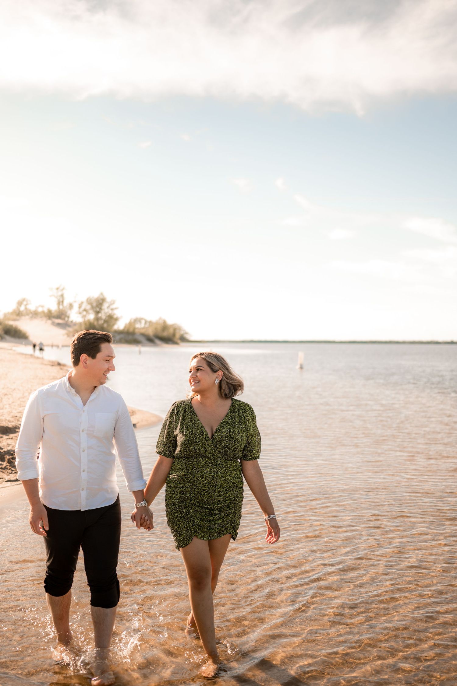 Happy couple holding hands and walking along a sunny shallow beach shoreline at golden hour.