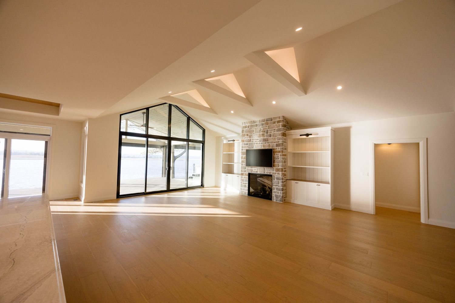 Empty living room with vaulted ceiling, large arched window, stone fireplace, built-in shelves, and hardwood floors.