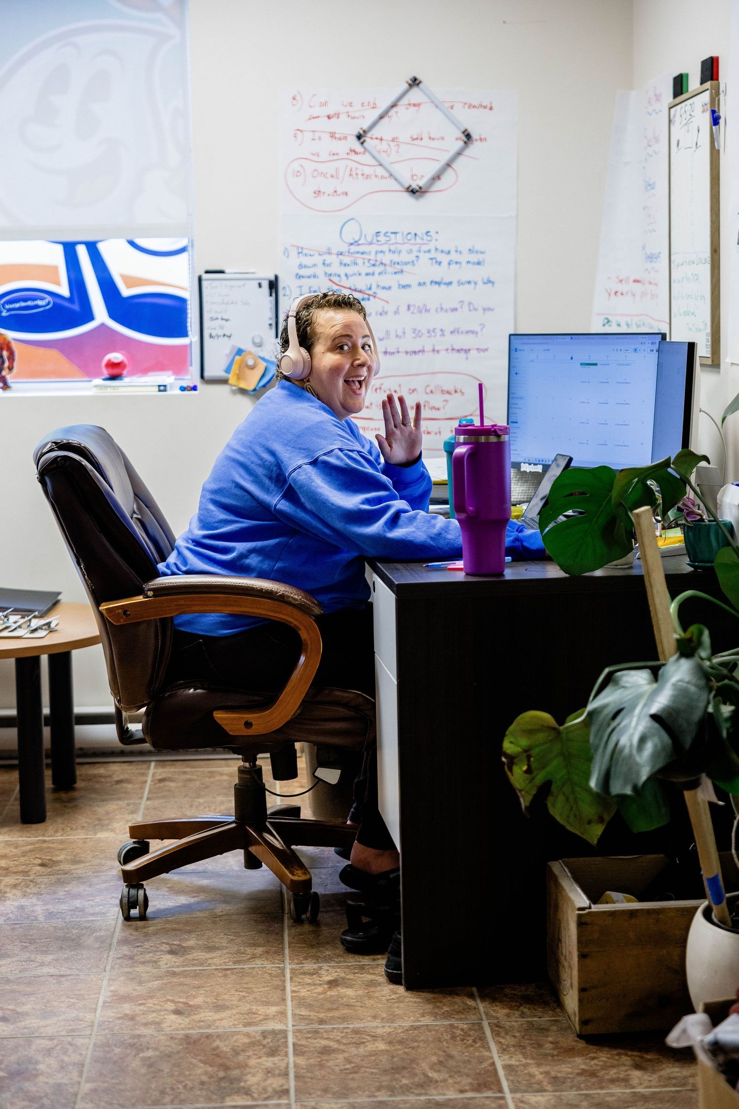 Friendly office staff member wearing headset waving at camera while working at desk.