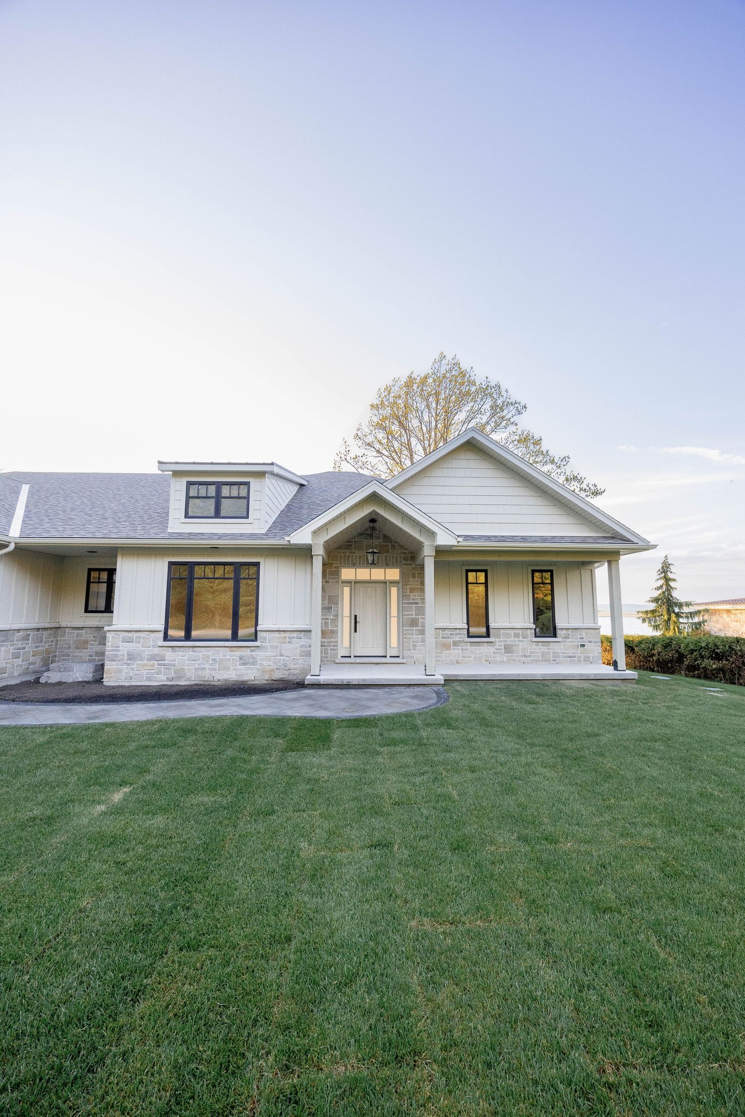 Modern white farmhouse with stone facade, black windows, and lush green lawn at dusk.