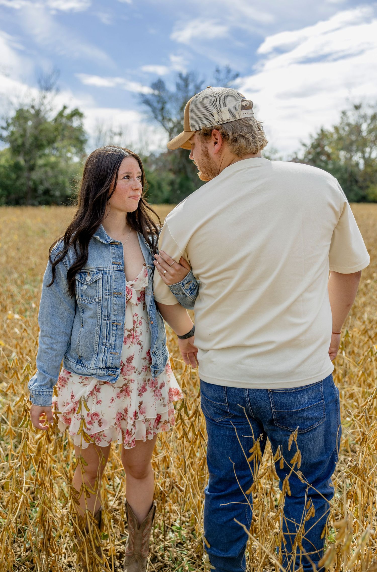A couple stands in a golden wheat field, the woman in a floral dress and denim jacket, the man in a cap and jeans.