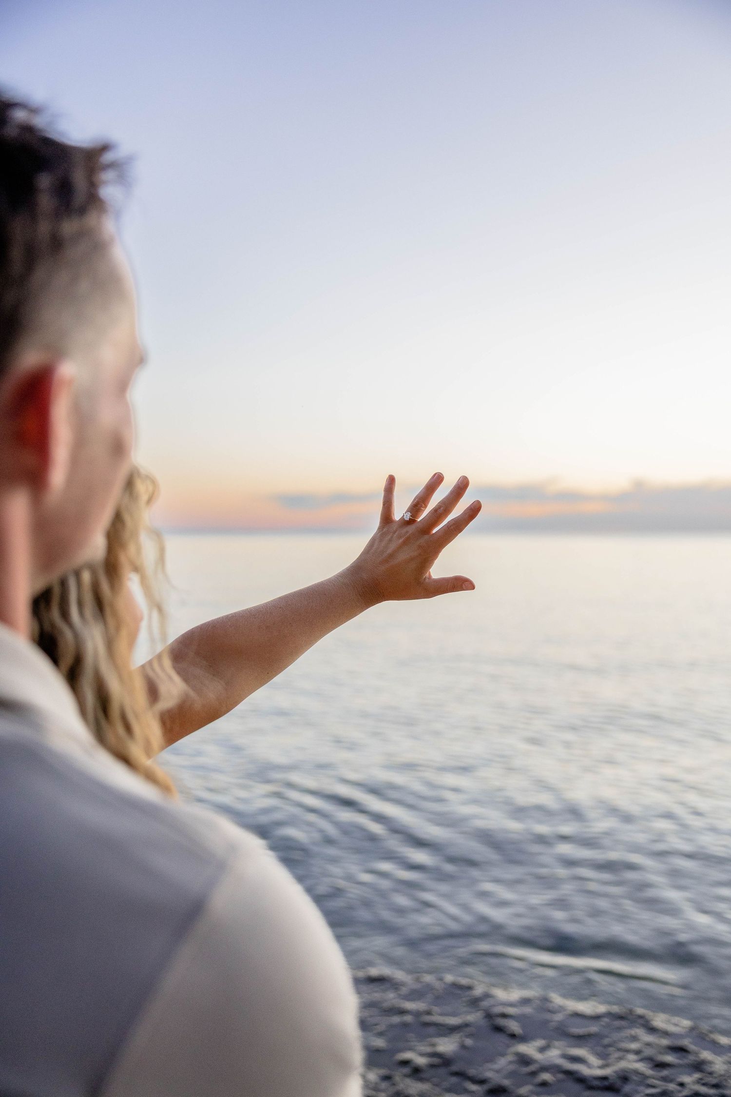 Person reaching hand toward calm ocean horizon at sunset, standing near rocky shoreline in soft evening light.