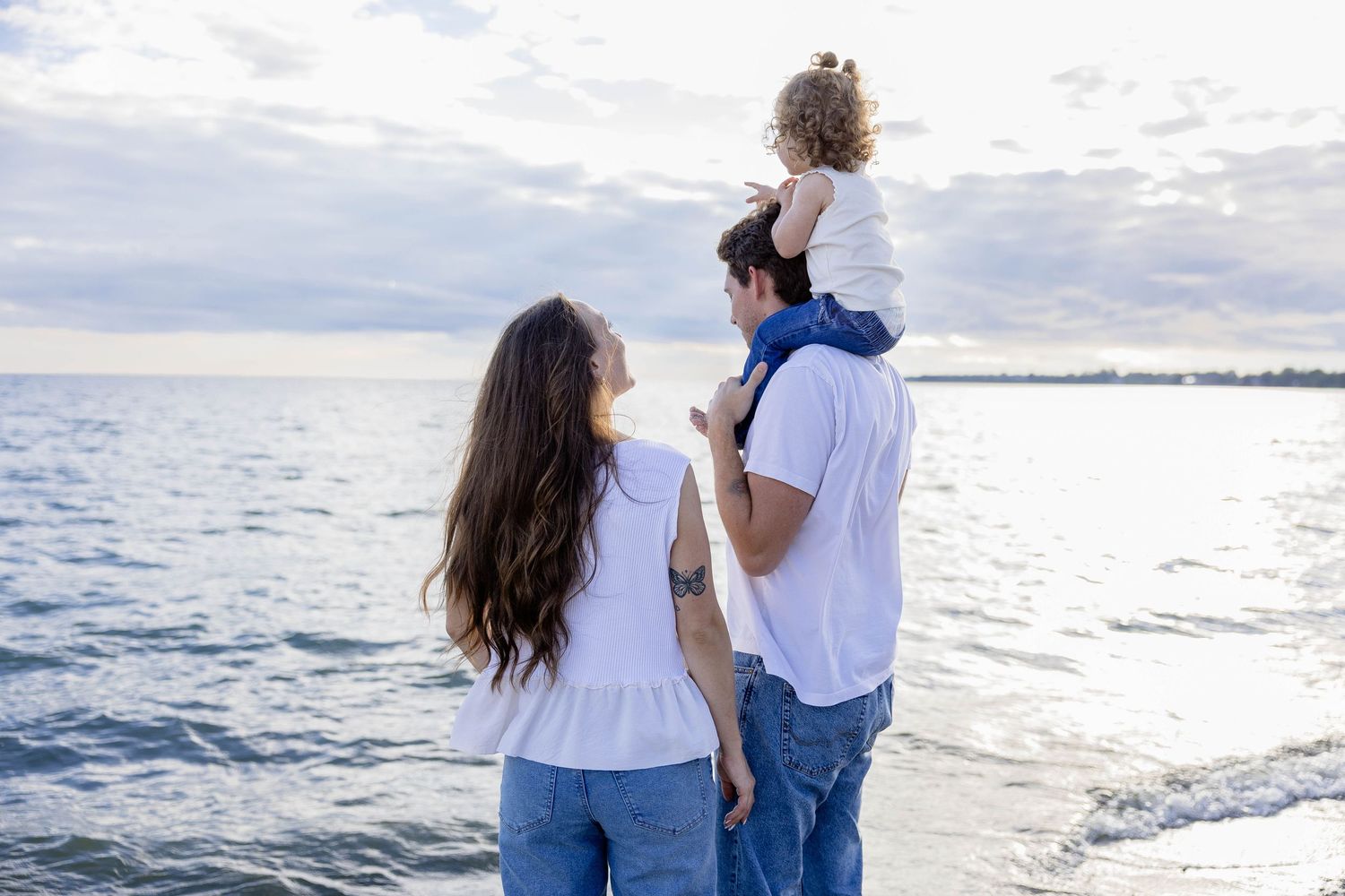 Family of three enjoying a serene beach sunset, parents in white shirts and jeans with child sitting on father's shoulders.