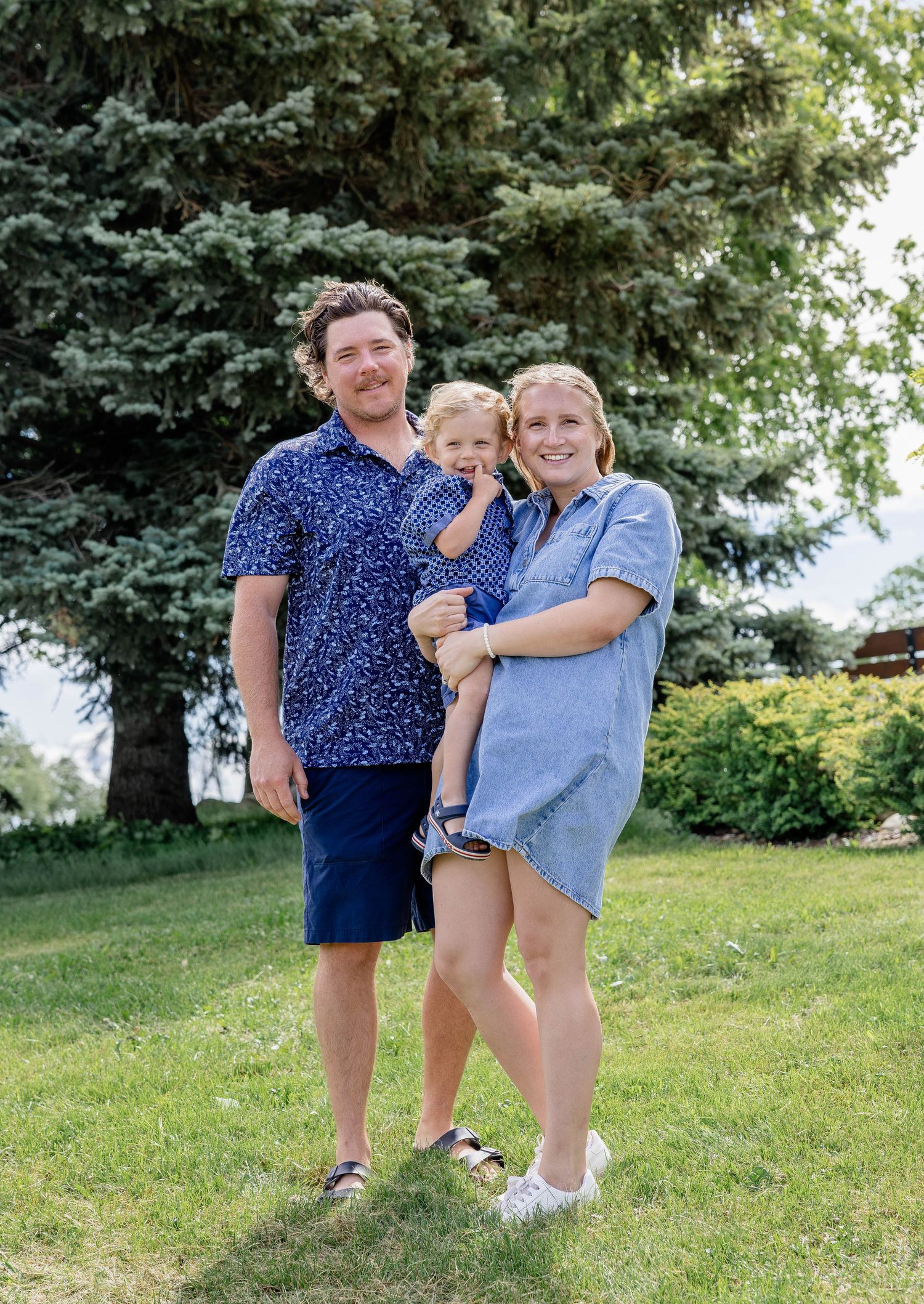 Happy young family with toddler posing outdoors on green lawn with evergreen trees in background on sunny day.