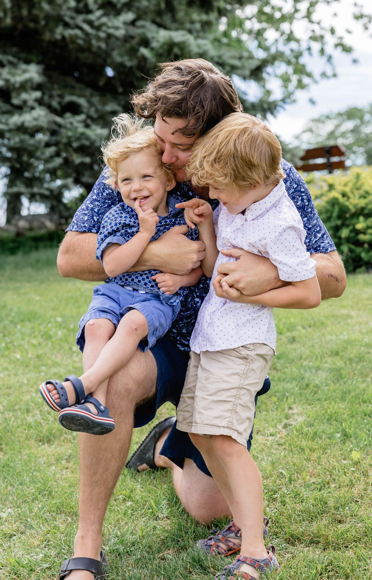A father kneels on grass hugging his two young blonde sons outdoors near green trees on a sunny day.