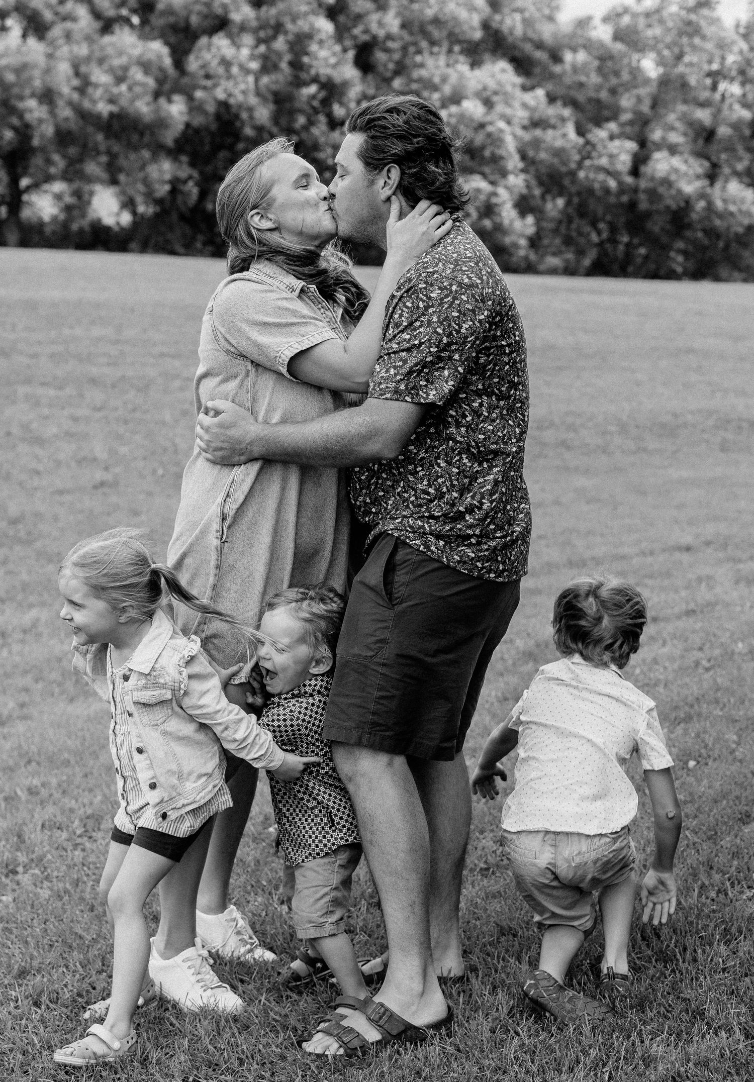 Black and white photo of a couple kissing in a park surrounded by young children on a grassy field.