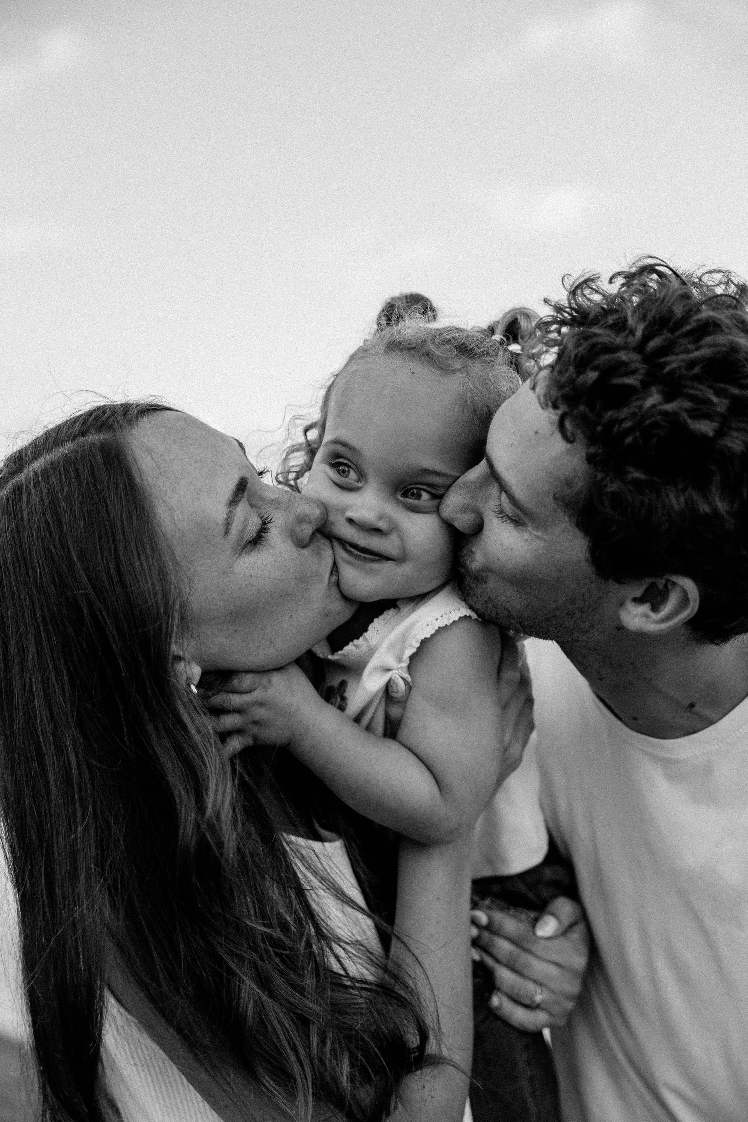 Black and white photo of two parents kissing their smiling baby on the cheeks outdoors.