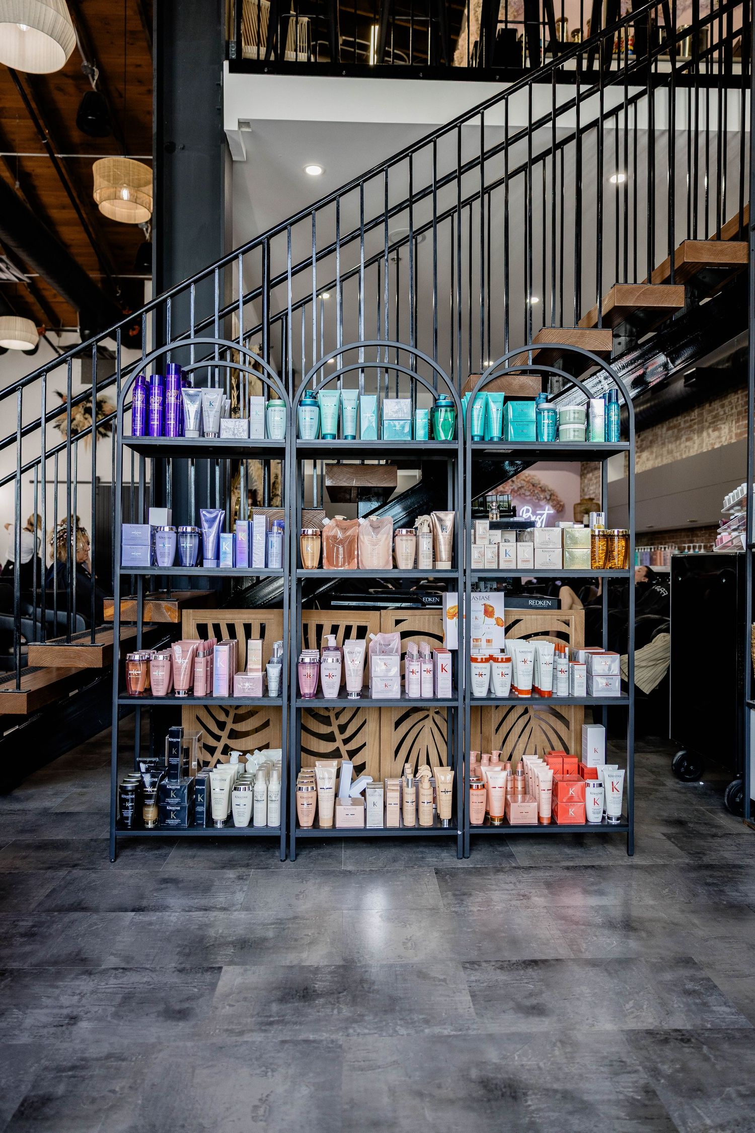 Metal retail shelving unit displaying colorful hair care products in a modern salon with industrial staircase.
