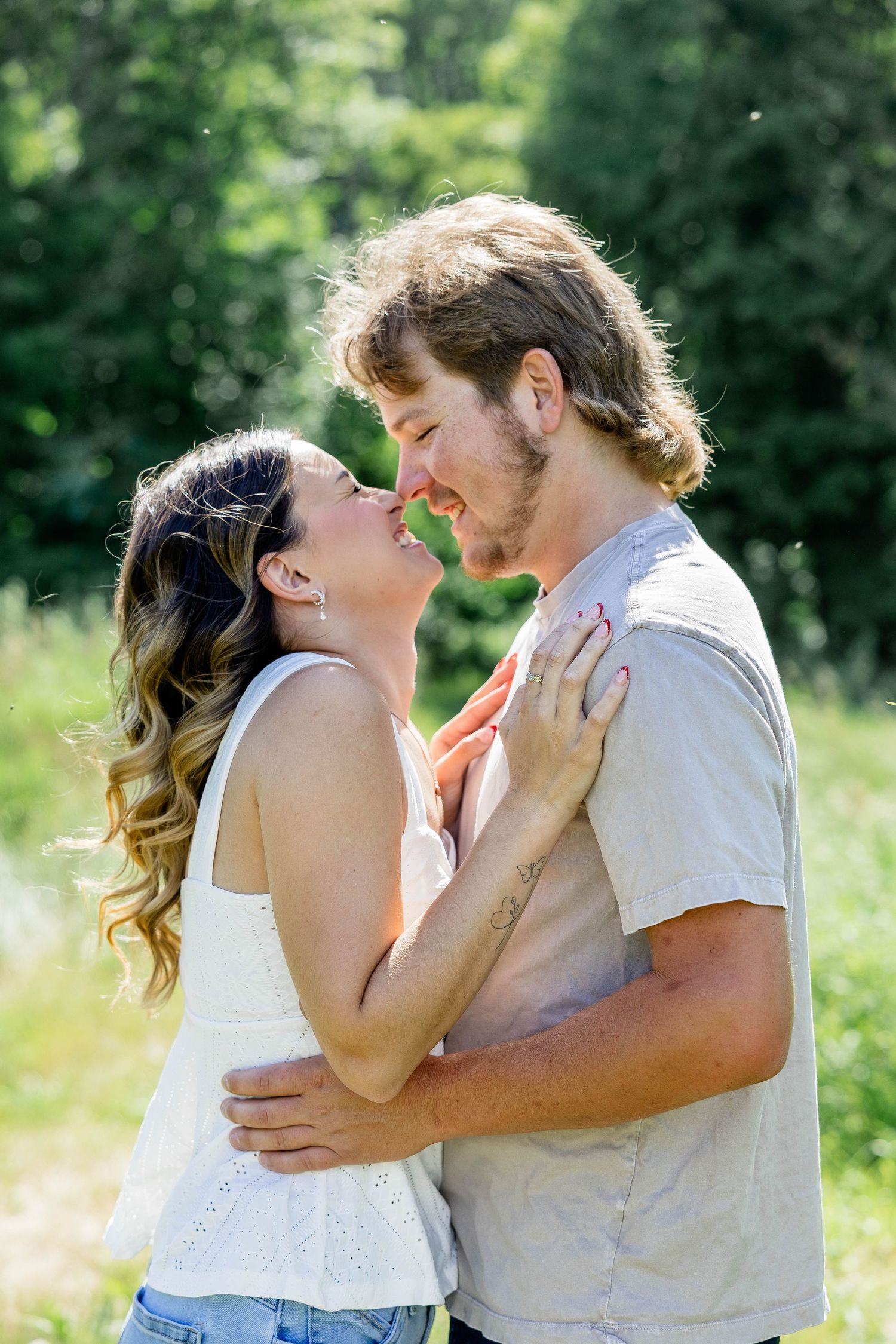 A couple embraces and smiles at each other in a sunny green outdoor setting, the woman in a white top and the man in a gray tee.