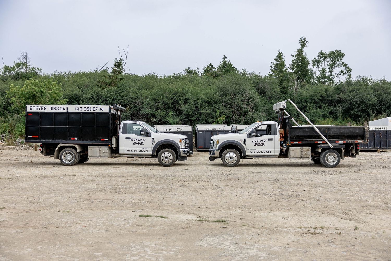 Two black dump trucks parked on gravel lot with company logos, surrounded by lush green trees on a cloudy day.
