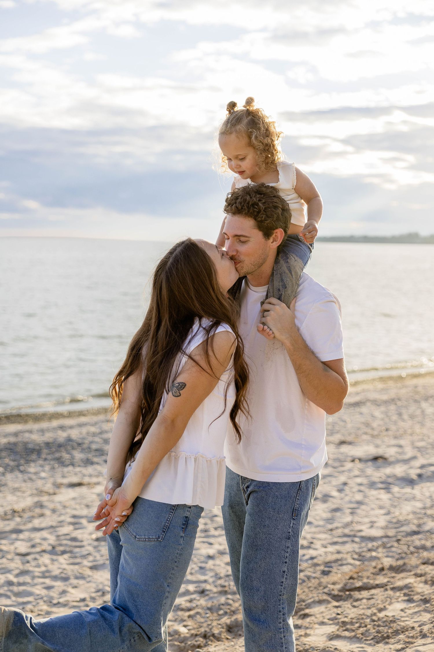 A young family enjoys a beach sunset, with dad carrying toddler on shoulders while mom kisses him on the cheek.