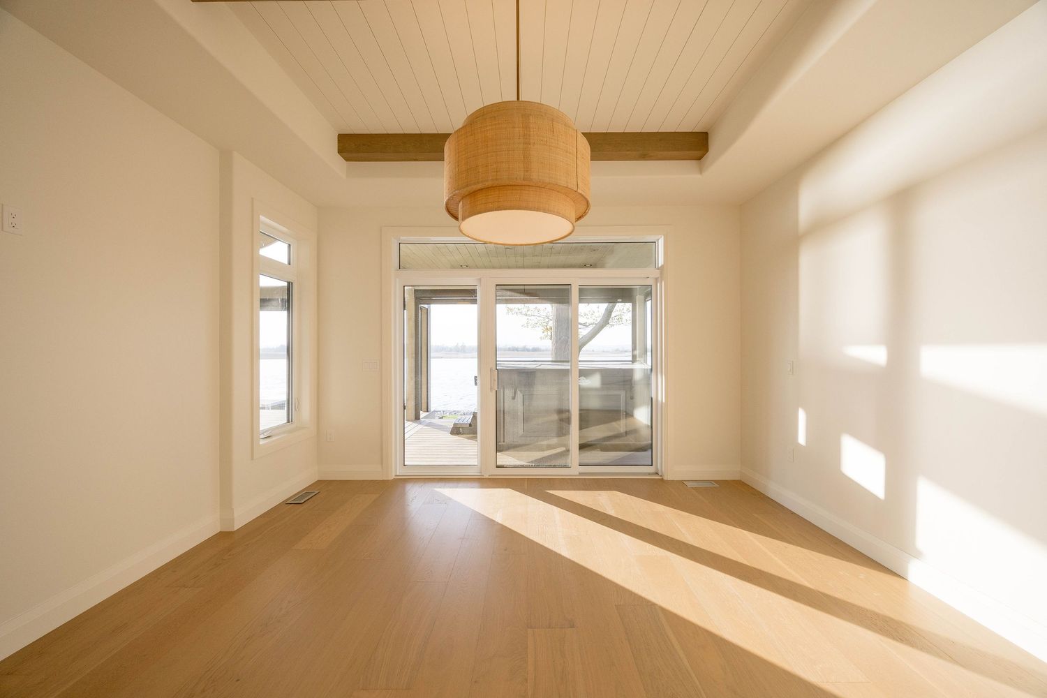 Bright empty room with beamed ceiling, woven pendant light, hardwood floors, and sliding glass doors.