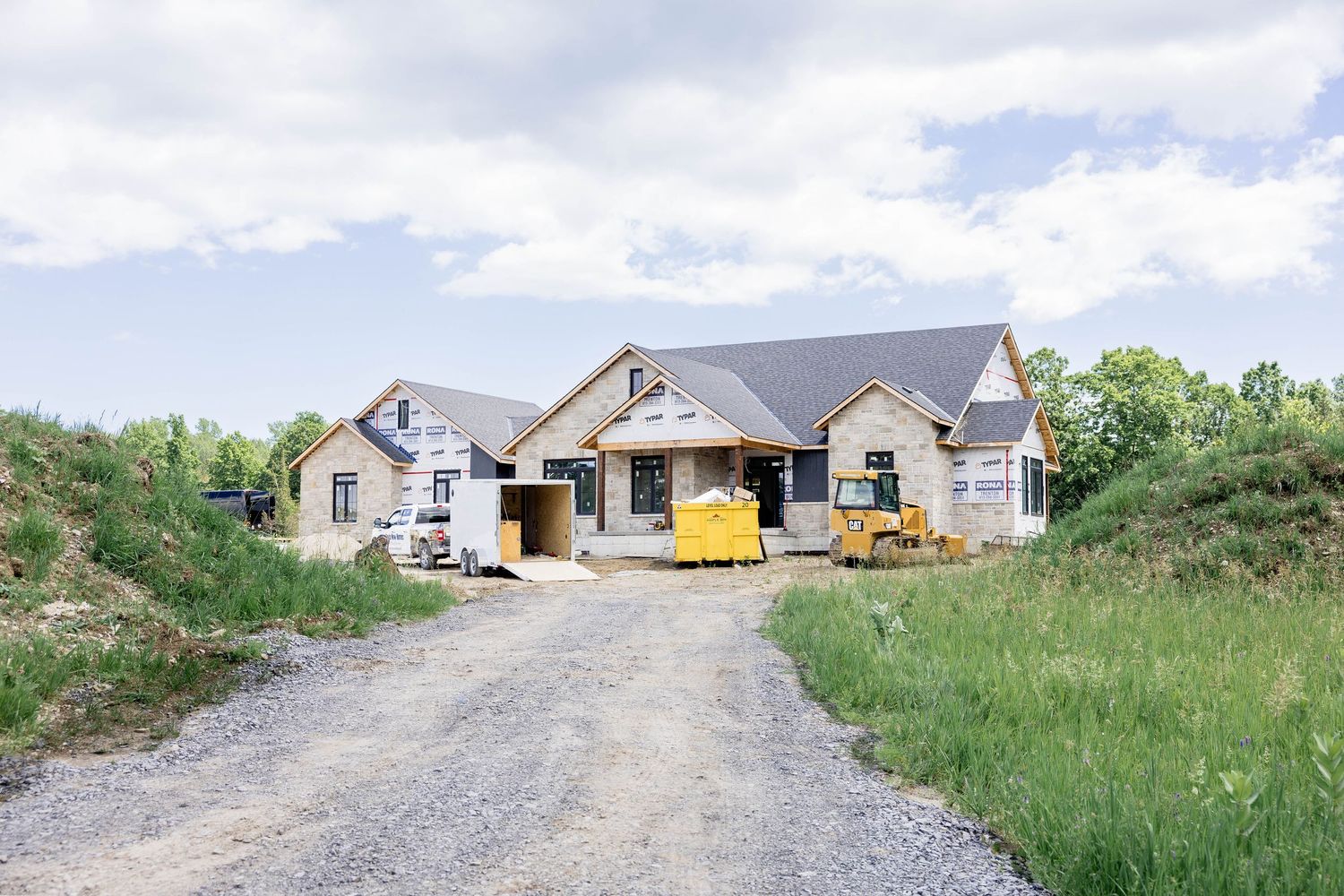 New home construction on gravel driveway with yellow dumpster and surrounding green landscape.