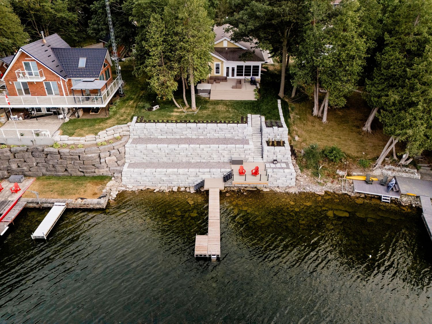 Aerial view of a lakeside property with a stone retaining wall, dock, and cottage surrounded by trees.