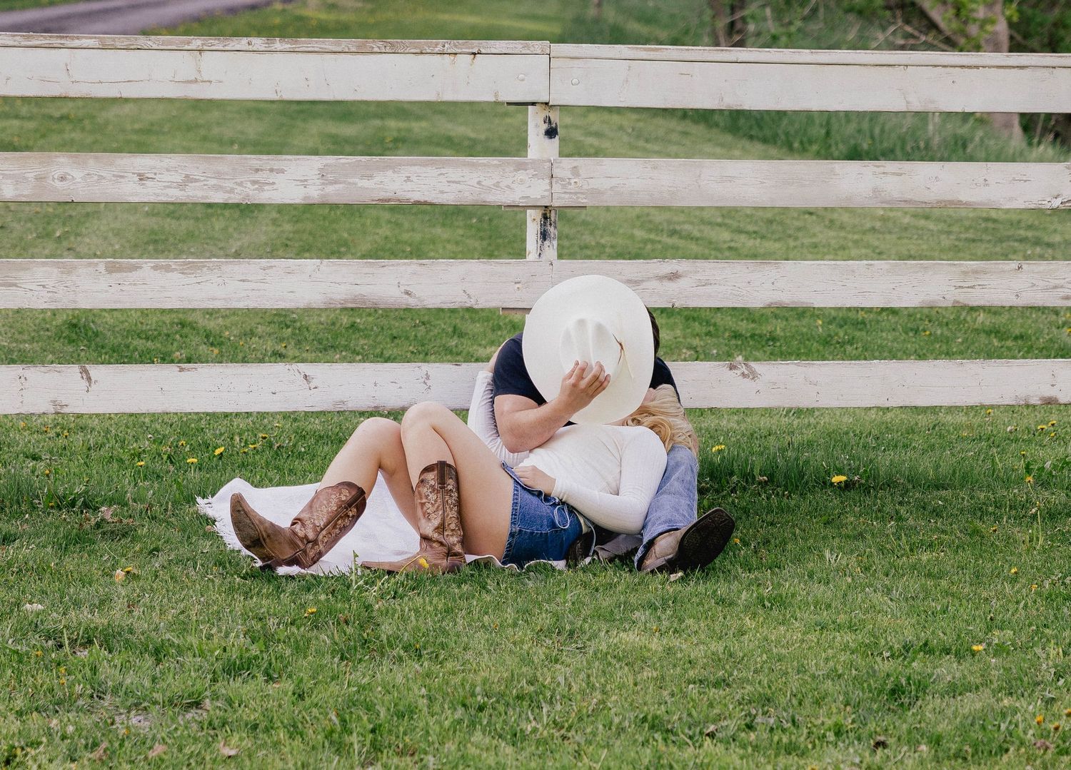 Couple kissing on grass by white fence, cowboy hat covering faces, western engagement photo.
