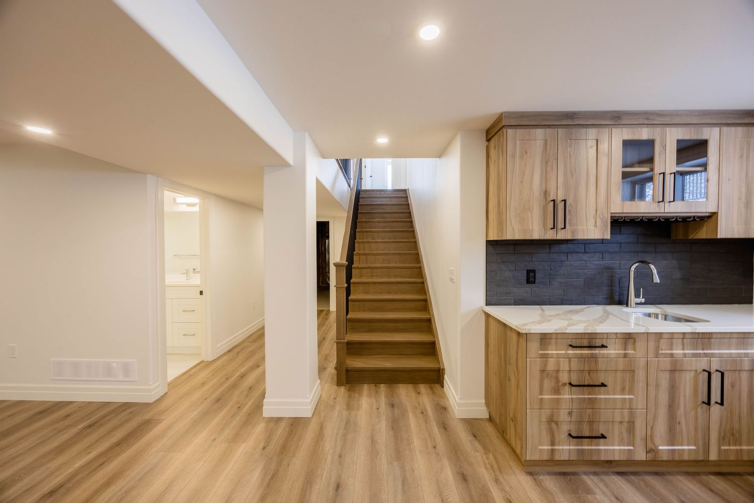 Modern basement wet bar with wood cabinetry, quartz countertops, and open staircase with vinyl flooring.