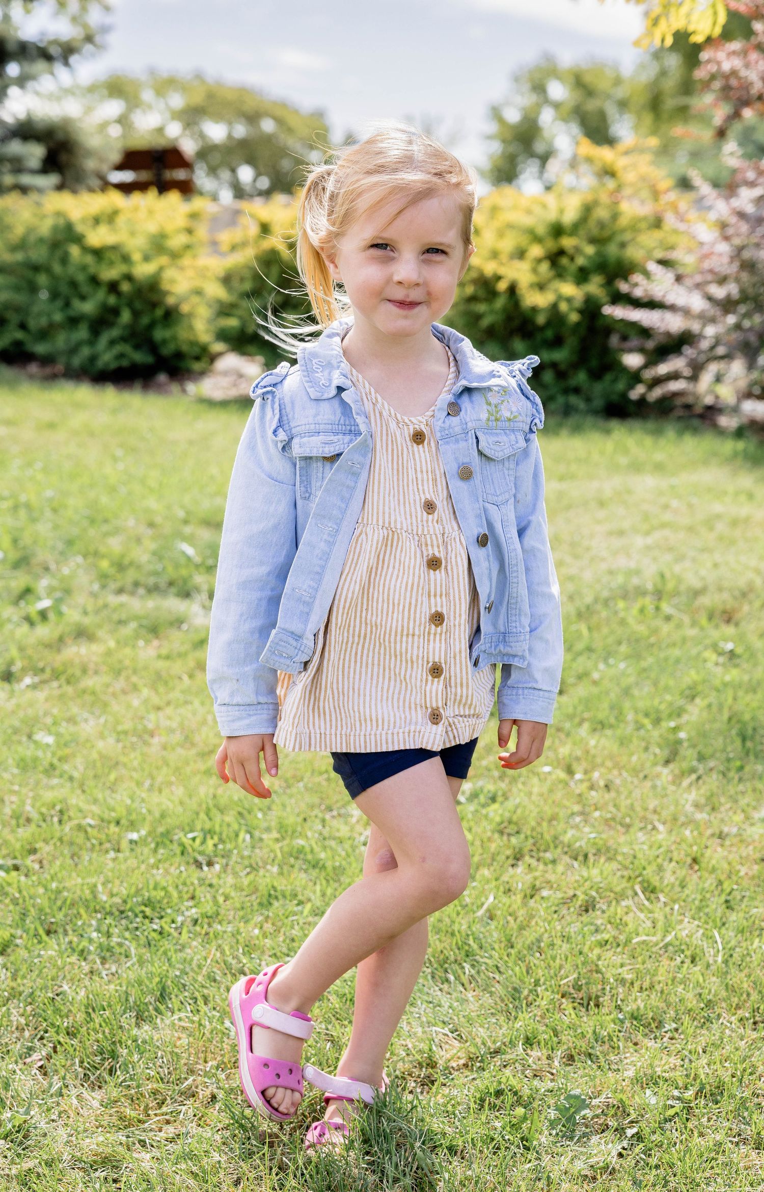 Smiling toddler girl in denim jacket, striped dress, navy shorts, and pink sandals posing on green grass outdoors.