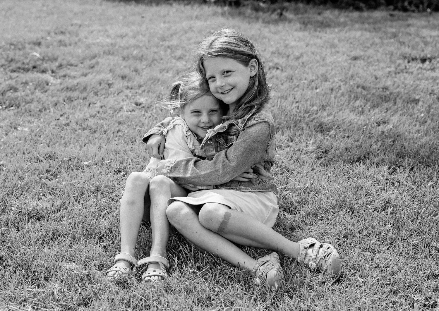Two young girls sitting on grass, sharing a warm hug, in a black and white outdoor photo.