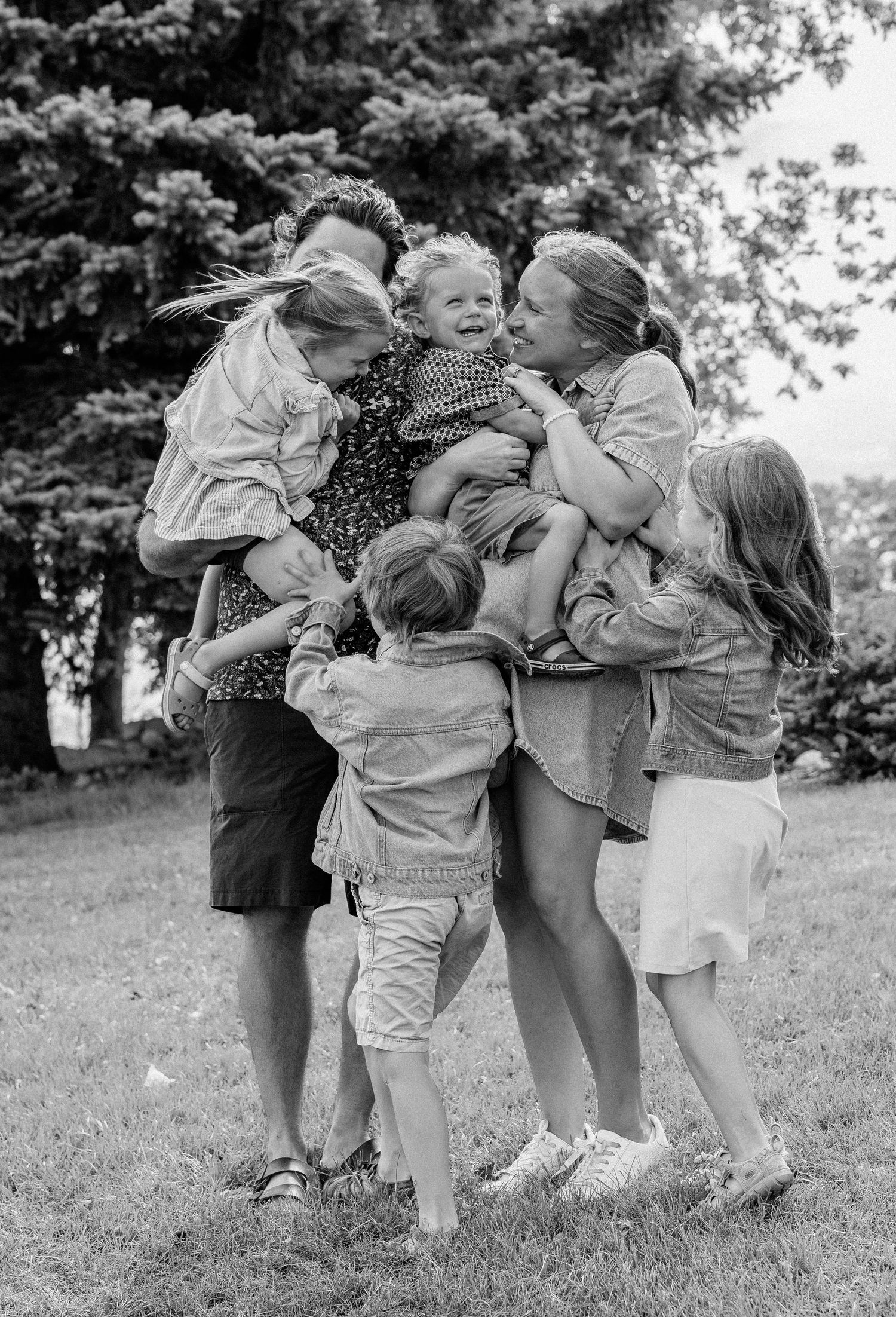 Black and white family photo of parents hugging and laughing with four young children outdoors on green grass.