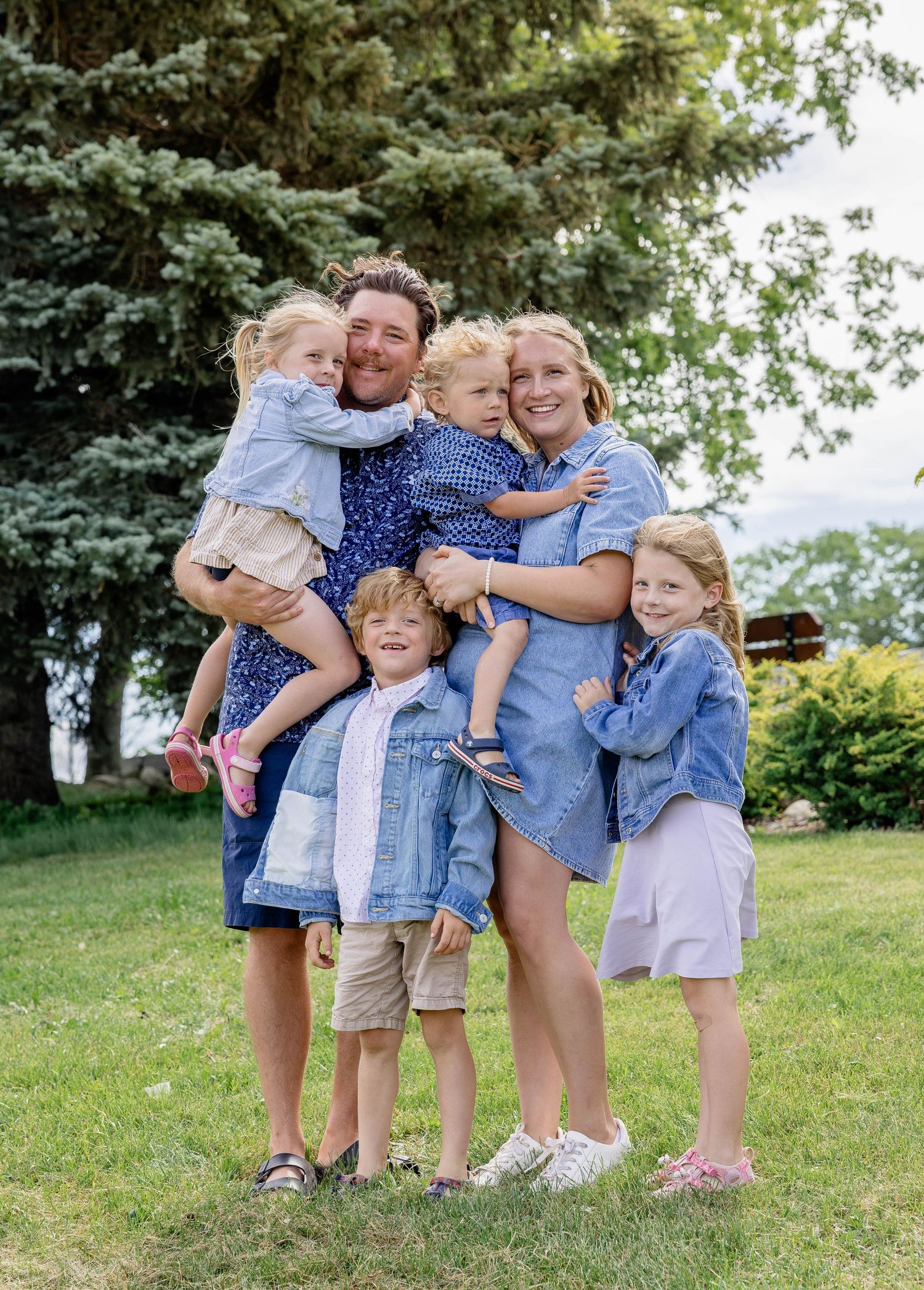 A happy family of six poses outdoors on green grass, smiling and hugging near a large evergreen tree on a sunny day.