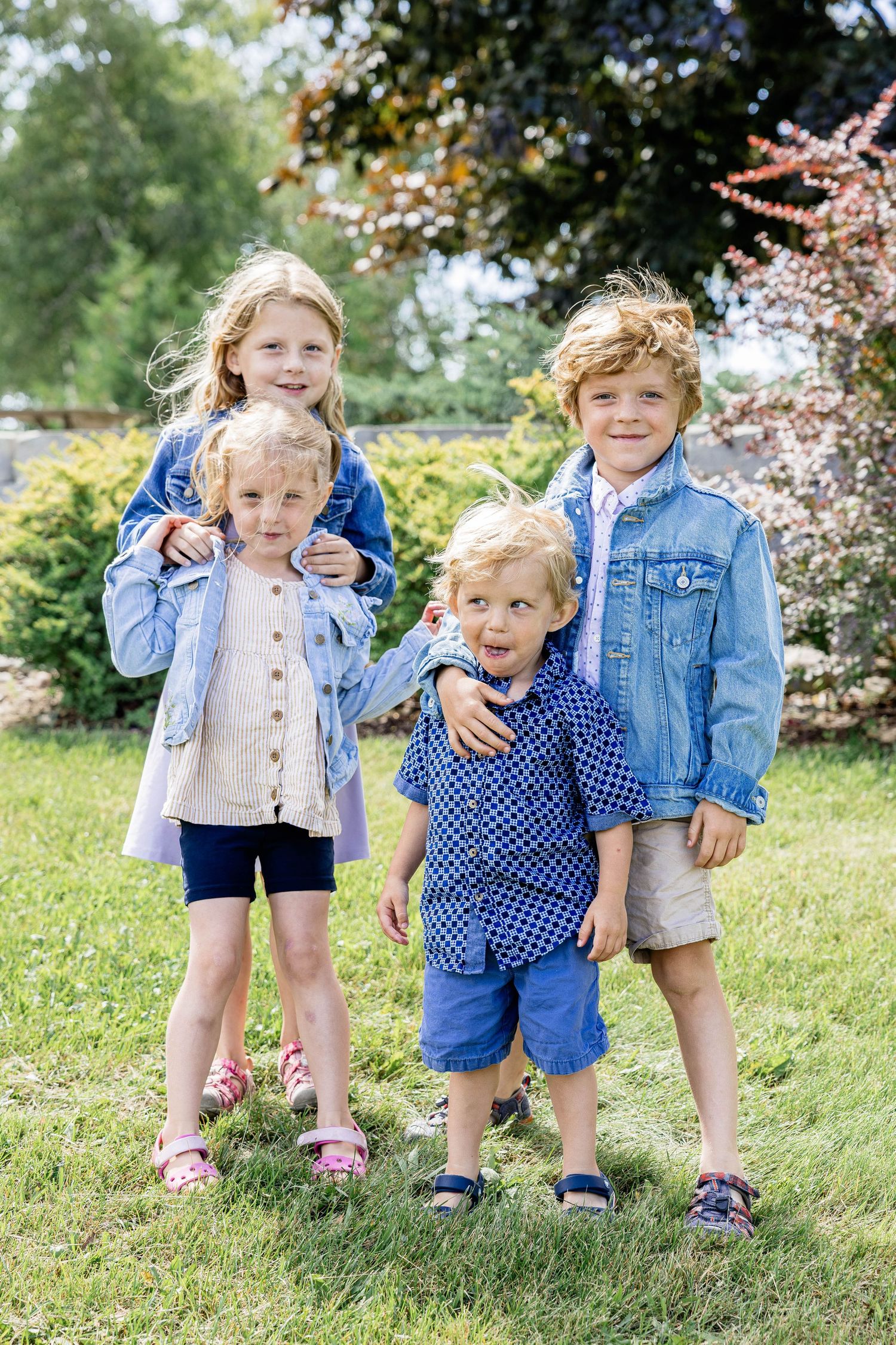Four young children in denim jackets pose together on green grass in a sunny garden setting.
