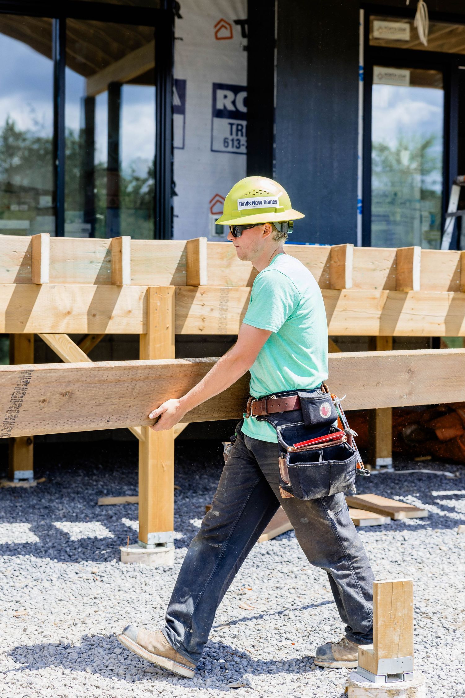 Construction worker in yellow hard hat carries lumber beam at residential building site.