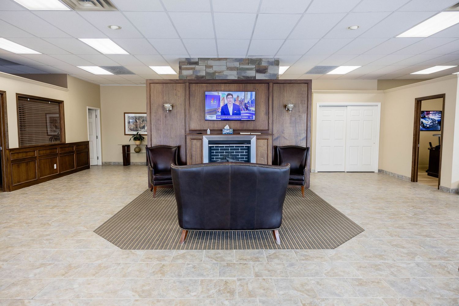Spacious car dealership waiting room with leather sofas, fireplace, and wall-mounted TV on tiled floors.