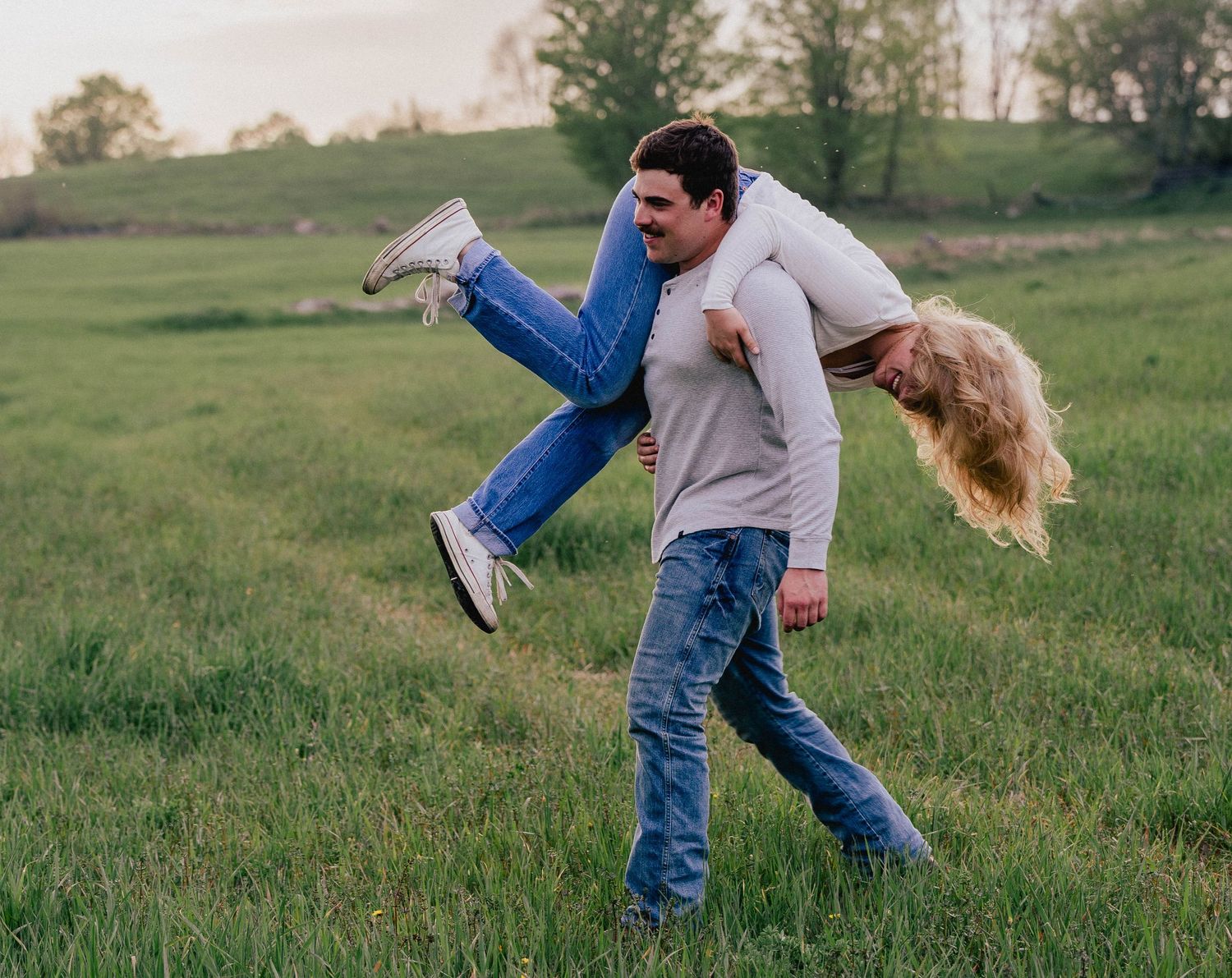 Man carrying woman over his shoulder in a green grassy field during a fun outdoor couples photo session.