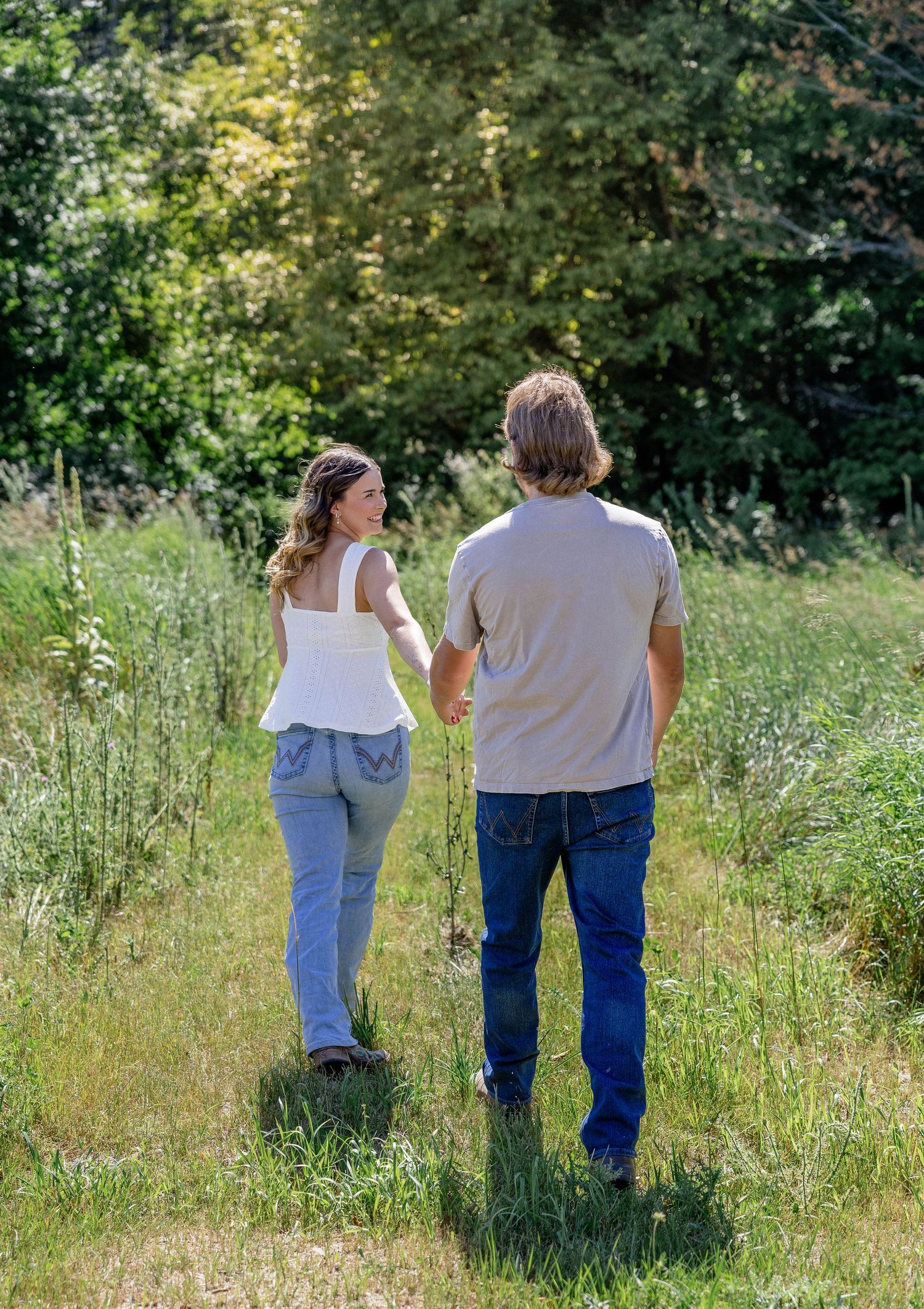 Couple holding hands walking through lush green meadow path surrounded by trees on a sunny day.
