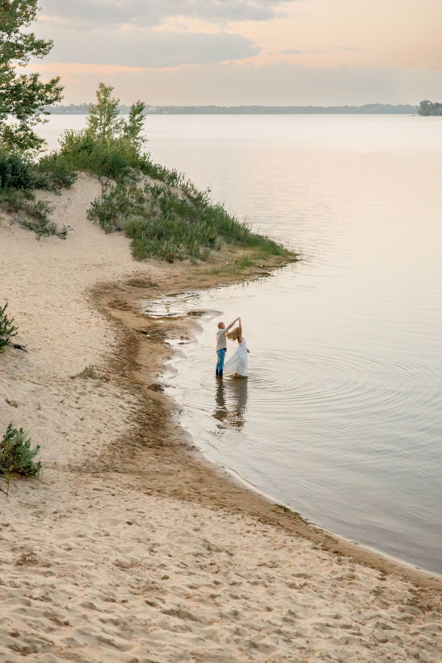 Couple dancing in shallow water at a sandy beach shore during a golden sunset evening.