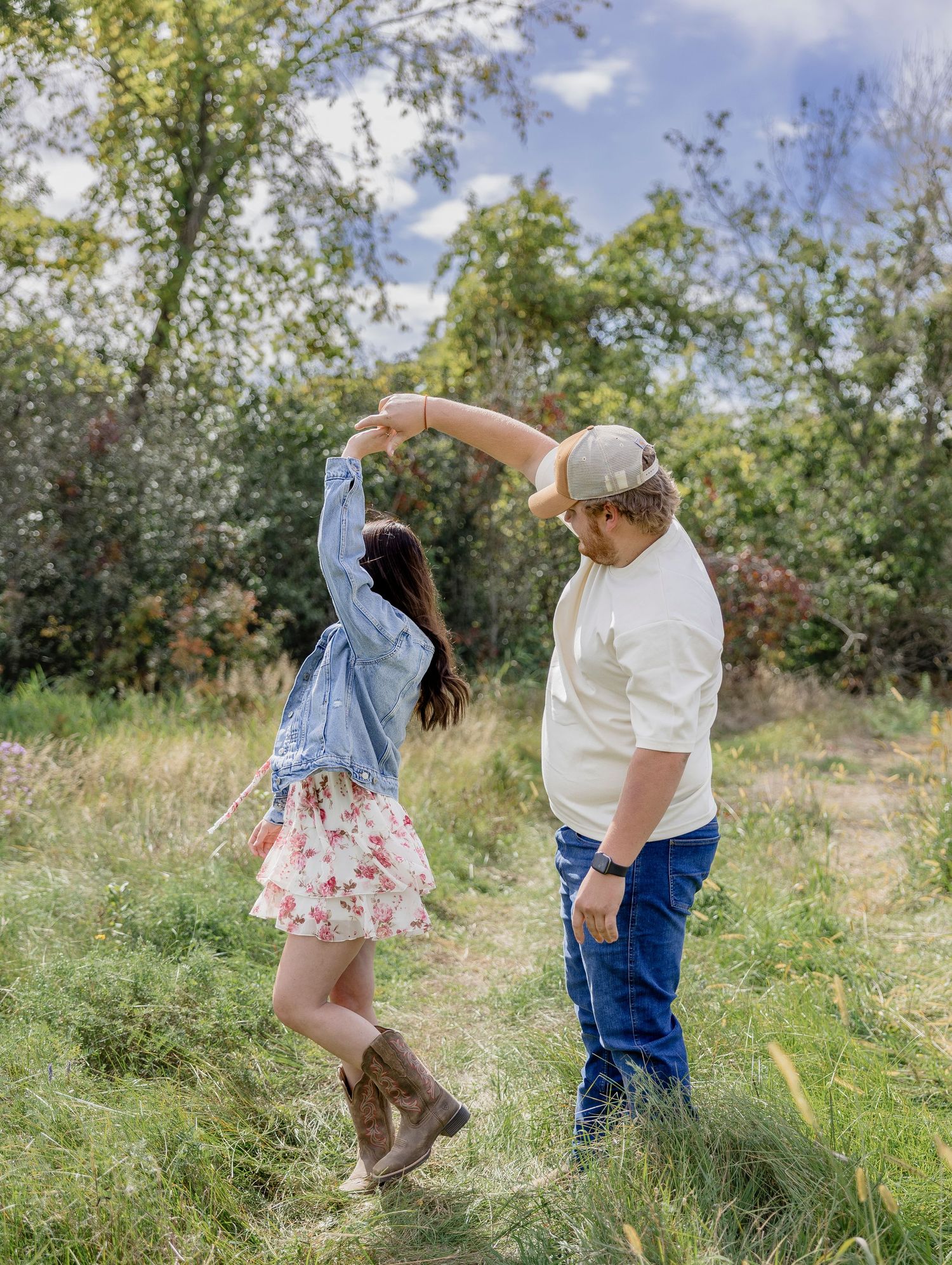 Couple dancing outdoors in a green meadow, woman in floral skirt and denim jacket, man in white shirt and cap.