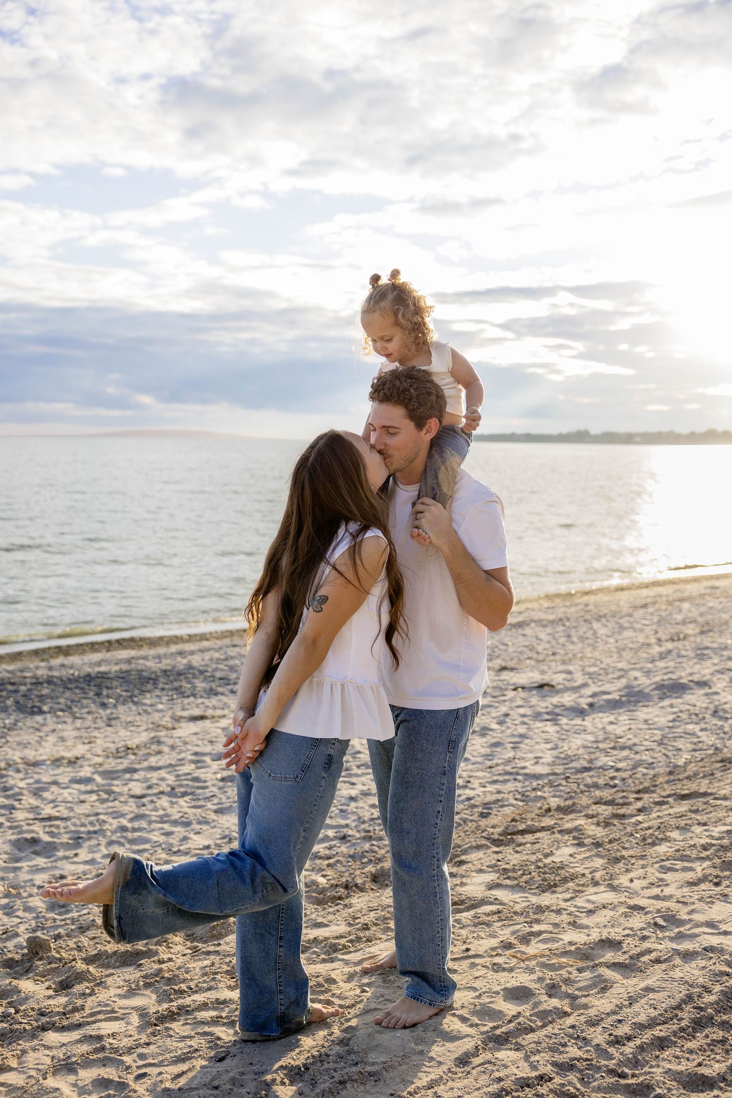A young family shares a kiss on a sandy beach at sunset, with a toddler sitting on dad's shoulders.
