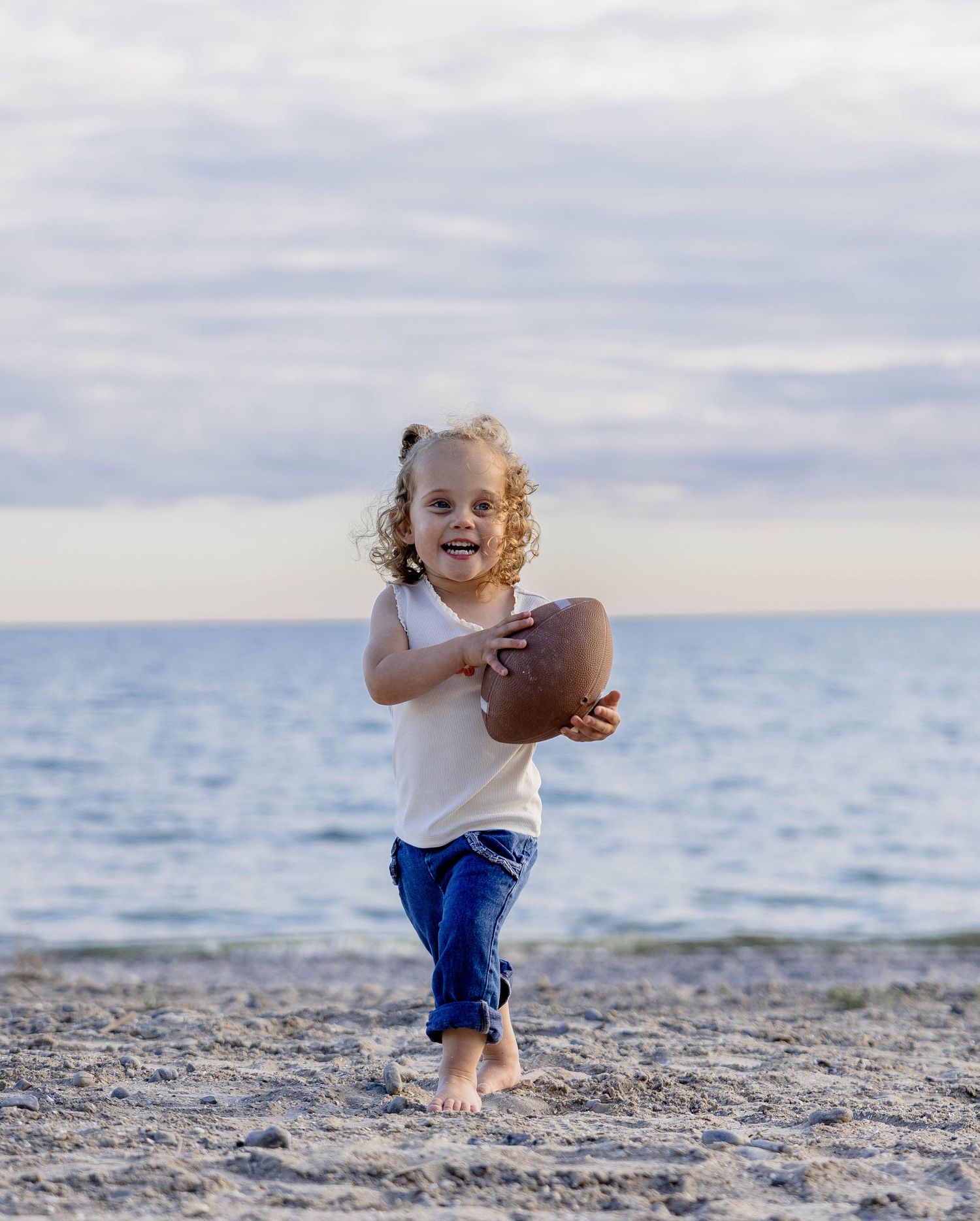 A happy toddler girl runs barefoot on a sandy beach holding a football, wearing a white tank top and blue jeans.